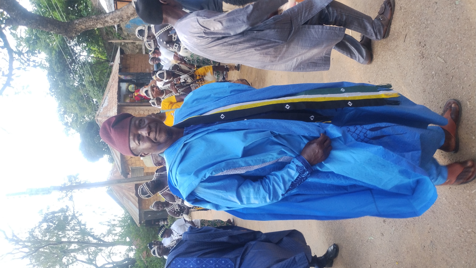 In the city of Jos, Nigeria, a man dressed in traditional blue attire, including a buba, sokoto, and a burgundy cap, stands in the foreground. He wears a colorful sash around his neck with stripes of black, yellow, green, and white. His feet are clad in brown leather sandals. Behind him, several other individuals in traditional clothing and headwear are visible. Some are adorned with beaded necklaces and headdresses, suggesting a cultural ceremony or festival. In the background, simple mud-brick structures with corrugated iron roofs are present, characteristic of some residential areas in Jos. The ground is a dirt path. The lighting suggests it is daytime, with bright sunlight filtering through the trees overhead.