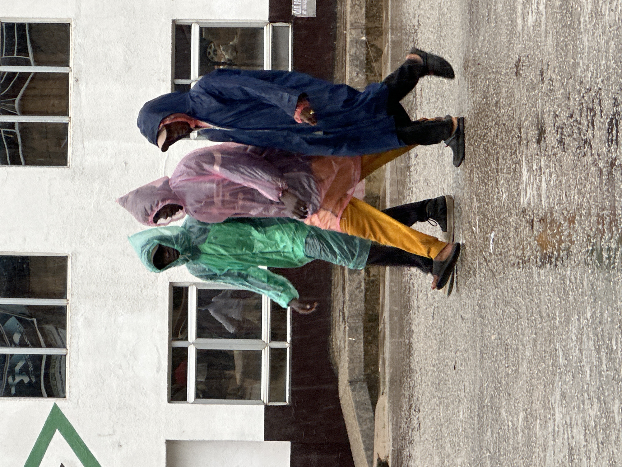 Three individuals are walking from left to right in the rain on a wet paved surface, possibly in Bukuru, Nigeria. The person on the left wears a translucent green hooded raincoat. The middle person is dressed in a translucent pink hooded raincoat over a light-colored shirt and yellow trousers. The person on the right is wearing a dark blue hooded raincoat over a pink garment and black trousers. The background features a white building with dark brown trim and windows. Reflections of people can be seen in the windows. The ground is covered in a thin layer of water, indicating ongoing rainfall, and shows ripples and reflections.