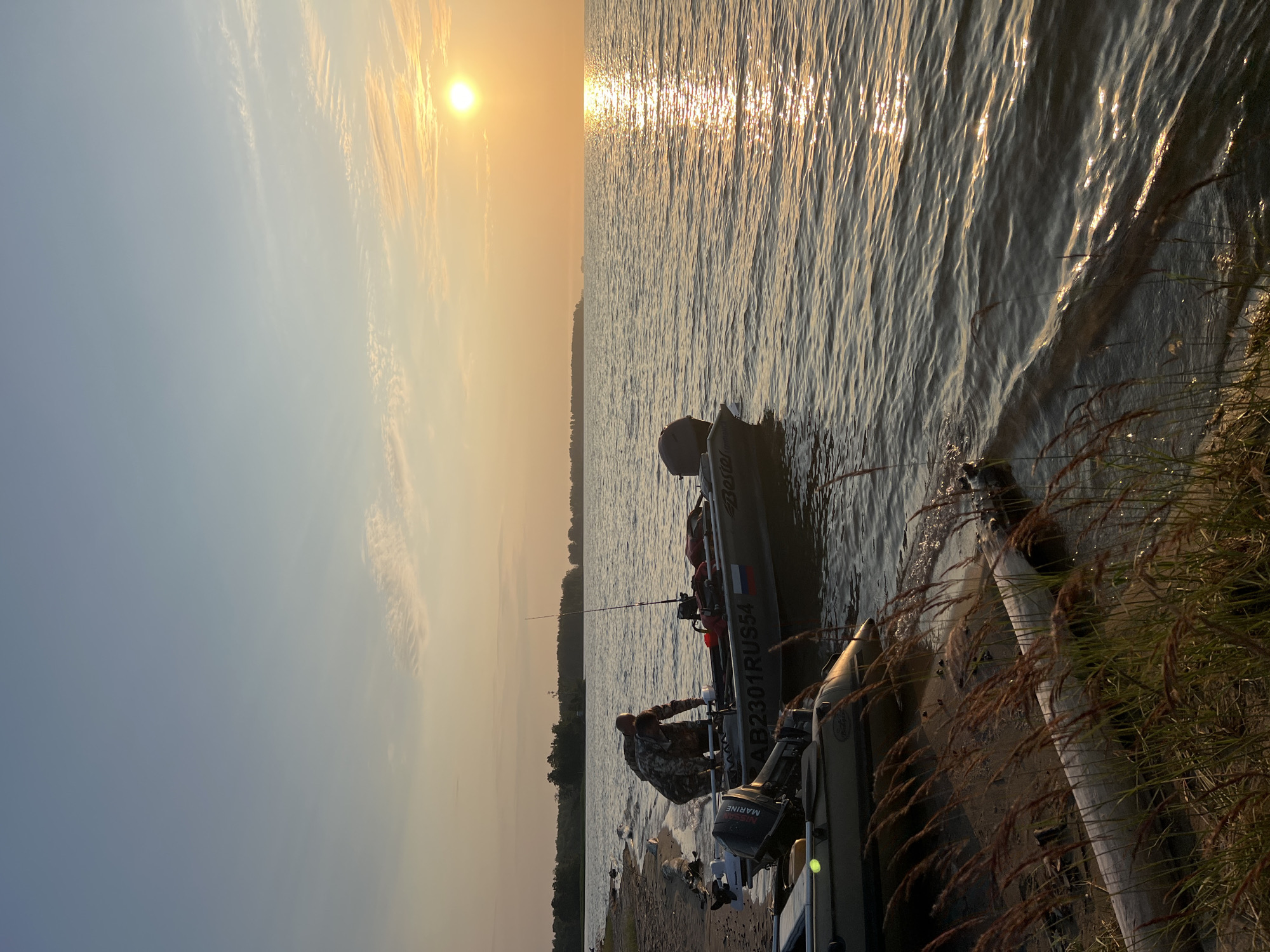 The image depicts a serene sunset over a body of water, likely Lake Borovoe, in Borovoe, Russia. The sky transitions from a pale blue at the top to a warm orange near the horizon where the sun is setting. Wispy clouds are scattered across the sky, catching the golden light. The water reflects the sunset, creating a shimmering path of light.
On the shore, two boats are partially submerged in the shallow water. One boat, a larger aluminum vessel with an outboard motor, has a person in camouflage attire standing in it, appearing to be preparing for an activity. The boat is marked with "AB2301RUS54" and a Russian flag. Another, smaller inflatable boat is docked next to it. The shoreline is sandy and littered with driftwood and tall, dry grass. Trees form a dark line in the distance along the opposite shore. The overall atmosphere is peaceful and suggests an outdoor recreational setting.