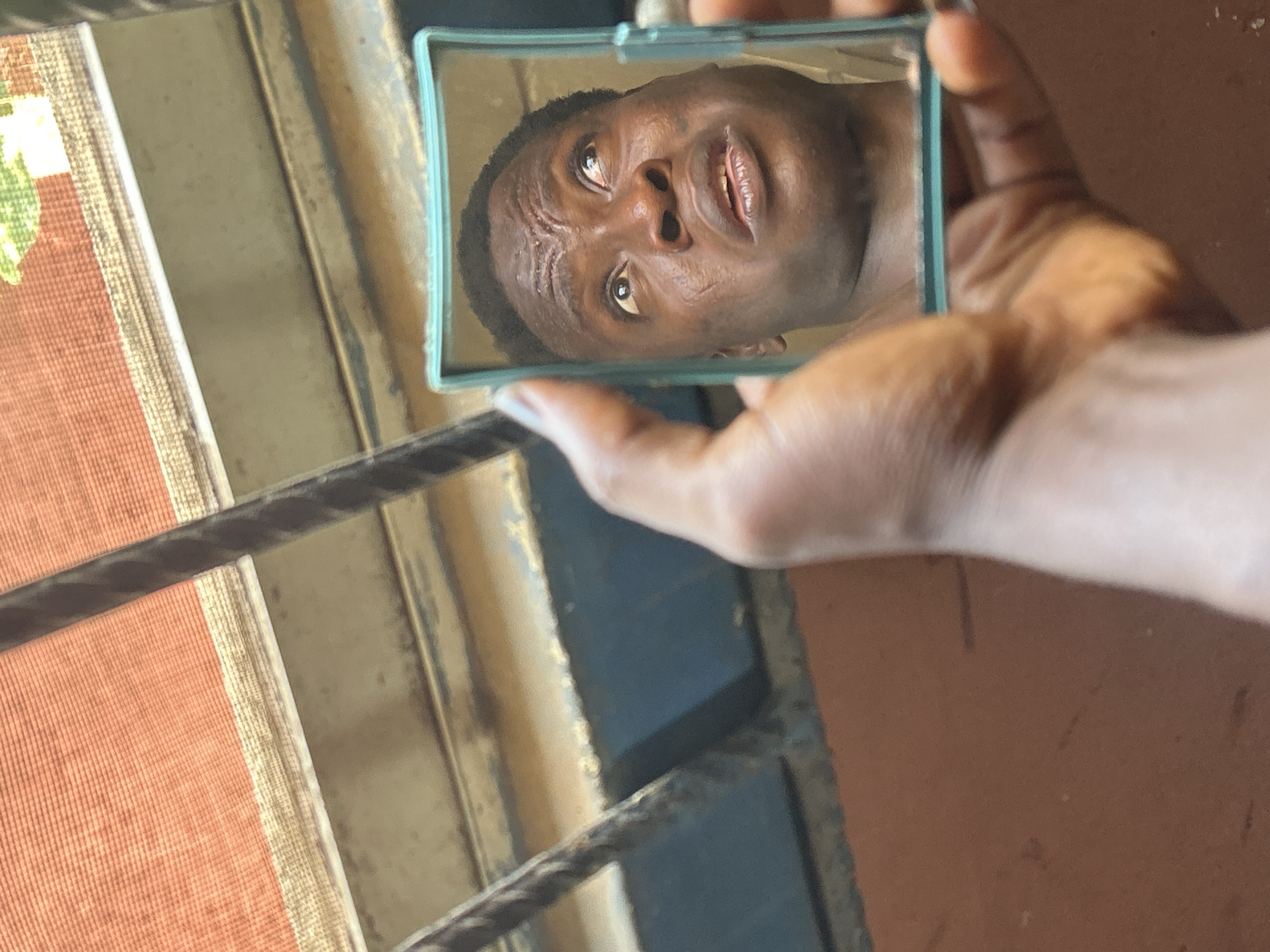 A dark-skinned person holds a small, rectangular mirror with a light blue frame, reflecting their face. The reflected face, likely an adult male, gazes upwards and slightly to the right, exhibiting a curious, concentrated, or slightly concerned expression with furrowed brows and a partially open mouth. The hand holding the mirror is also dark-skinned.
The setting appears to be indoors, near a window or opening, illuminated by bright natural daylight, indicating it is daytime. A thick, dark, horizontal bar, possibly a window grill or railing, spans the middle left of the frame. To the left of this bar, a textured reddish-orange material with lighter vertical stripes, resembling blinds or a curtain, is visible. To the right and in the corners, there is a warm-toned, possibly terracotta-colored, wall or surface. No visible text is present in the image.