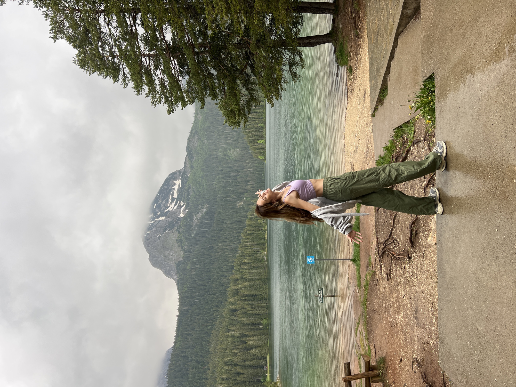 The image, when correctly oriented, presents a scenic landscape from Pitomine, Montenegro, featuring a person by a lake surrounded by mountains and dense forests under an overcast sky.
In the foreground, a young woman with long, dark brown hair stands on a dirt path next to the water. She is wearing a light purple tank top, olive green cargo pants, and grey and white athletic shoes. A grey hooded jacket is draped over her shoulders. She leans backward with her head tilted towards the sky and eyes closed, conveying a sense of serenity or enjoyment of the moment.
The middle ground is dominated by a tranquil, greenish-blue lake with subtle ripples on its surface. Beyond the lake, majestic mountains rise, covered extensively by evergreen forests. The upper reaches of the mountains show patches of snow and exposed rock, suggesting a rugged terrain. The sky is uniformly cloudy and grey, indicating overcast weather, and the ground near the lake appears damp.
To the right of the woman, the dirt path transitions into a concrete walkway or steps. Exposed tree roots are visible on the ground near the path. A large evergreen tree with abundant foliage hangs over the scene from the upper left.
Near the water's edge, two signs are visible. One is a blue square sign featuring a white circle with a symbol, likely indicating a prohibition such as "no fires" or "no camping." Below it, a wooden sign reads, "PLEASE KEEP IT CLEAN! LEAVE NO TRACE," promoting environmental responsibility. The overall atmosphere is one of calm and natural beauty.
