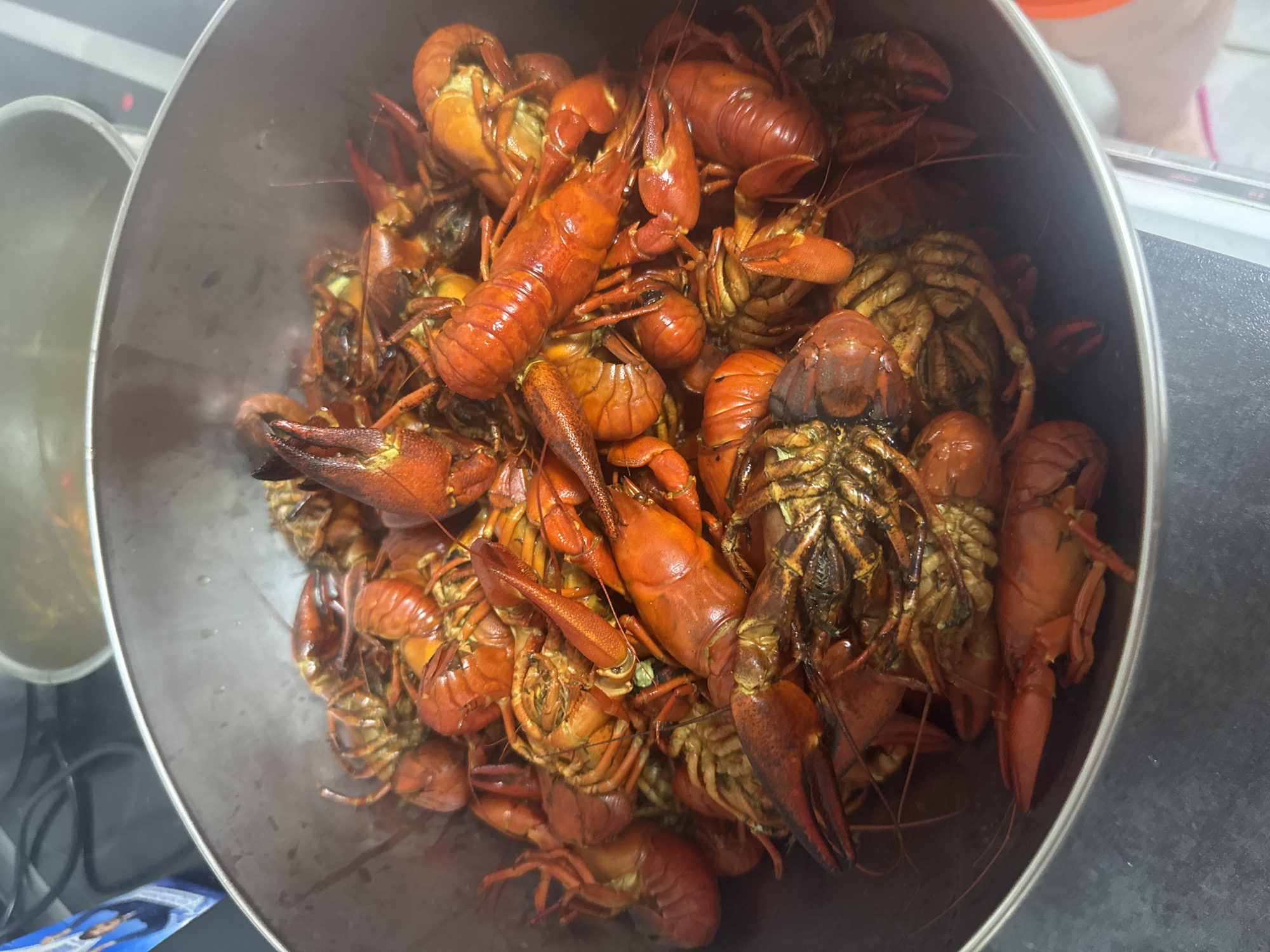 A large, silver metal bowl is filled to the brim with cooked crawfish. The crawfish are bright red and orange, with their shells partially curled. Some crawfish are stacked on top of others, and their claws are visible. The bowl is set on a dark gray countertop. In the background, a pot with a reflective surface and a partial view of a blue and white label are visible.