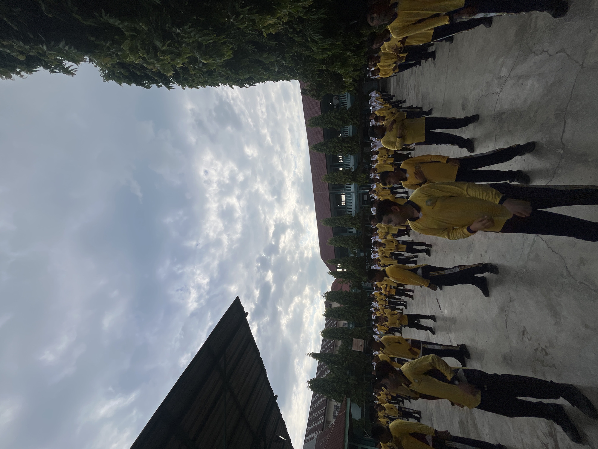 A low-angle shot captures a large group of young males, appearing to be students, in a coordinated physical activity outdoors. They are all dressed in matching yellow long-sleeved shirts and dark pants. The students are arranged in rows on a paved courtyard, some standing still with hands at their sides or on their hips, while others are mid-movement, suggesting a form of synchronized exercise or assembly.
The environment includes a covered walkway or stage structure on the left side of the frame, a row of tall, green trees in the background, and school buildings with red roofs behind the trees. The sky is overcast with visible clouds, indicating diffused natural light. The scene is set in Lhokseumawe, Indonesia, likely on the grounds of a school or educational institution, possibly during a morning assembly or physical education class.