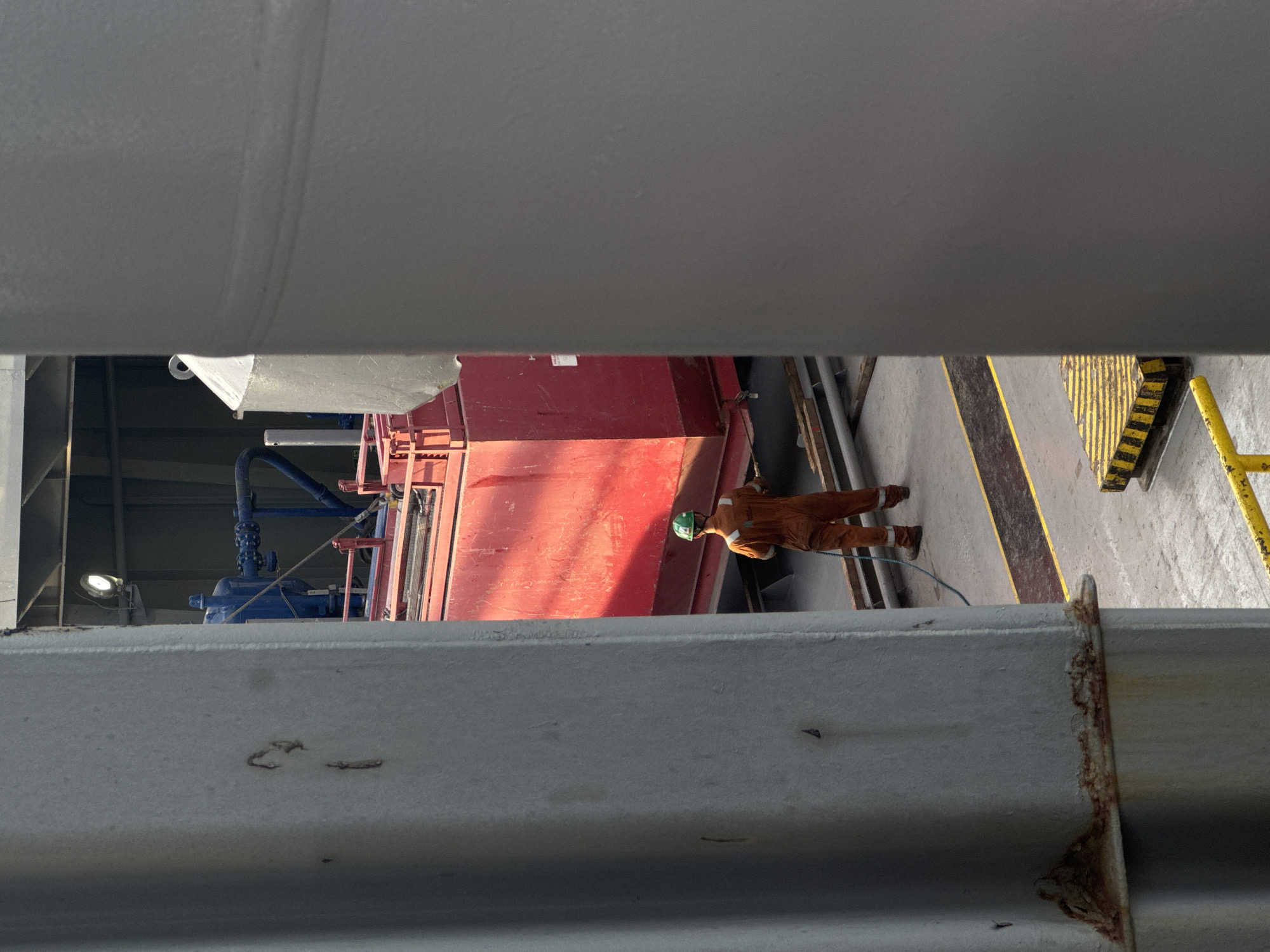 The image shows a person wearing an orange jumpsuit and a green hard hat, standing on a concrete floor. The person is facing away from the camera, towards a large red industrial container. The setting appears to be an industrial or port area, with metal structures and equipment visible. The lighting suggests it could be daytime, with sunlight casting a shadow. There are yellow and black striped safety markings on the floor, indicating a designated walkway or hazard area. The person appears to be working or inspecting the equipment. The overall impression is one of industrial activity.