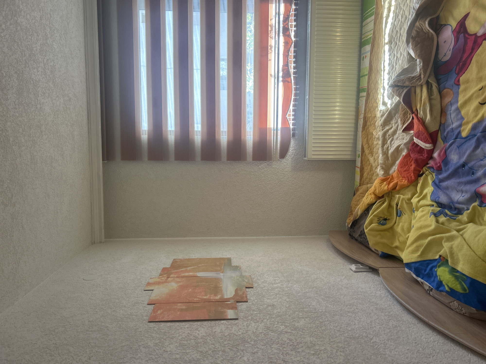 A sparsely furnished room with light-colored walls and carpet. In the foreground, there are several rectangular tiles stacked unevenly on the floor. Towards the right, a bed with colorful bedding is visible. A window is positioned in the center of the room with partially closed vertical blinds, revealing glimpses of the outside. A radiator is to the right of the window. The room appears to be located indoors. There is no visible indication of Zlatograd, Bulgaria.