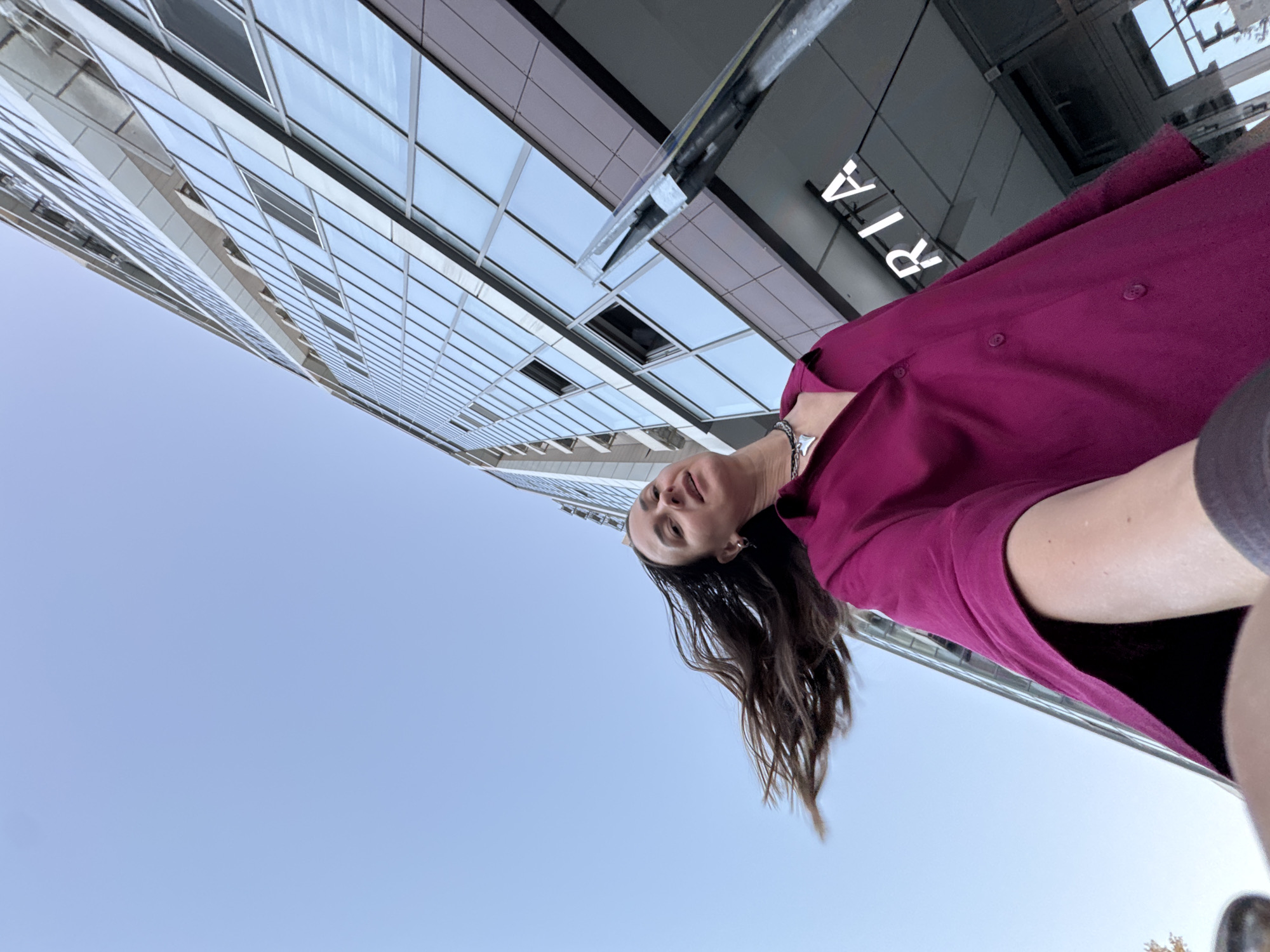 A low-angle, wide-shot photograph captures a woman standing outside a modern, glass-paneled building with her hair blowing in the wind. She is wearing a vibrant magenta shirt and a statement necklace. The building, located in the city of Sochi, Russia, appears to be an office building or a commercial establishment, with the partially visible illuminated sign spelling "RIA" suggesting it might be a media or news agency. The sky is a clear, pale blue. The composition, with its upward tilt and the woman's hair creating a dynamic element, suggests a candid moment or a selfie.