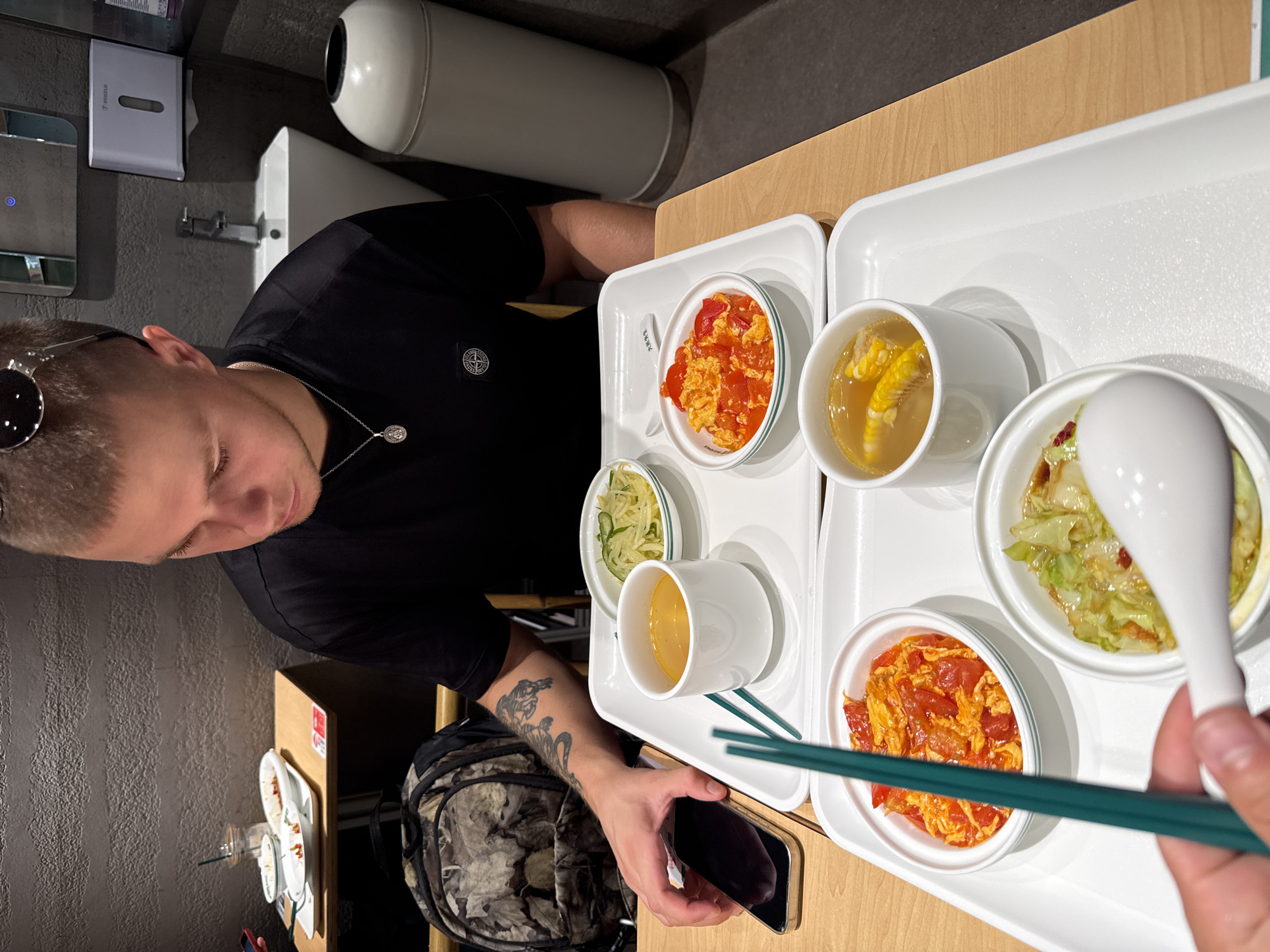 In Tianhe District, China, a young man with short blonde hair and sunglasses perched on his head sits at a table with a tray of food. He is wearing a black t-shirt and a necklace, and has tattoos on his left arm. He is looking down at his phone, which is placed on the tray. The tray contains several small bowls of food, including what appears to be scrambled eggs with tomatoes, a clear broth with corn, and a salad. A pair of green chopsticks rests on the tray, and a white spoon is partially visible in the foreground. In the background, a sink and a trash can are visible, suggesting the setting may be a restaurant or a similar public establishment. A camouflage backpack is placed on the floor next to the table.