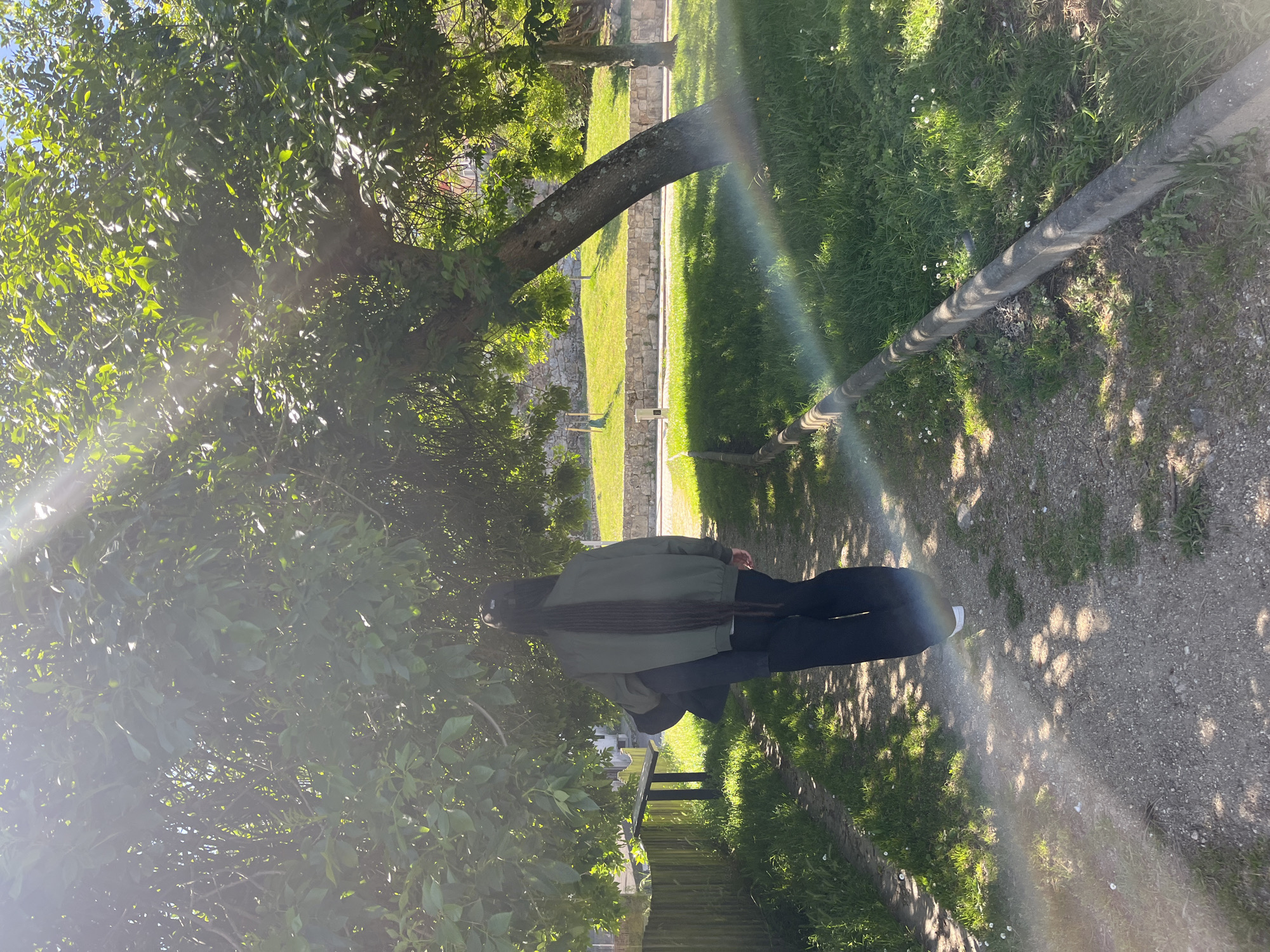 The photograph captures a view in A Coruña, Spain. The scene presents a person with long dreadlocks, clad in a green jacket and black pants, walking along a gravel path. The individual is partially obscured by the foliage of a tree, which branches out over the path and casts shadows. A wooden utility pole runs parallel to the path on the right. A strip of grass, interspersed with small white flowers, lies adjacent to the path. In the background, a stone wall is visible beyond another grass strip, and sunlight filters through the leaves of the tree, creating a rainbow effect in the center of the image.