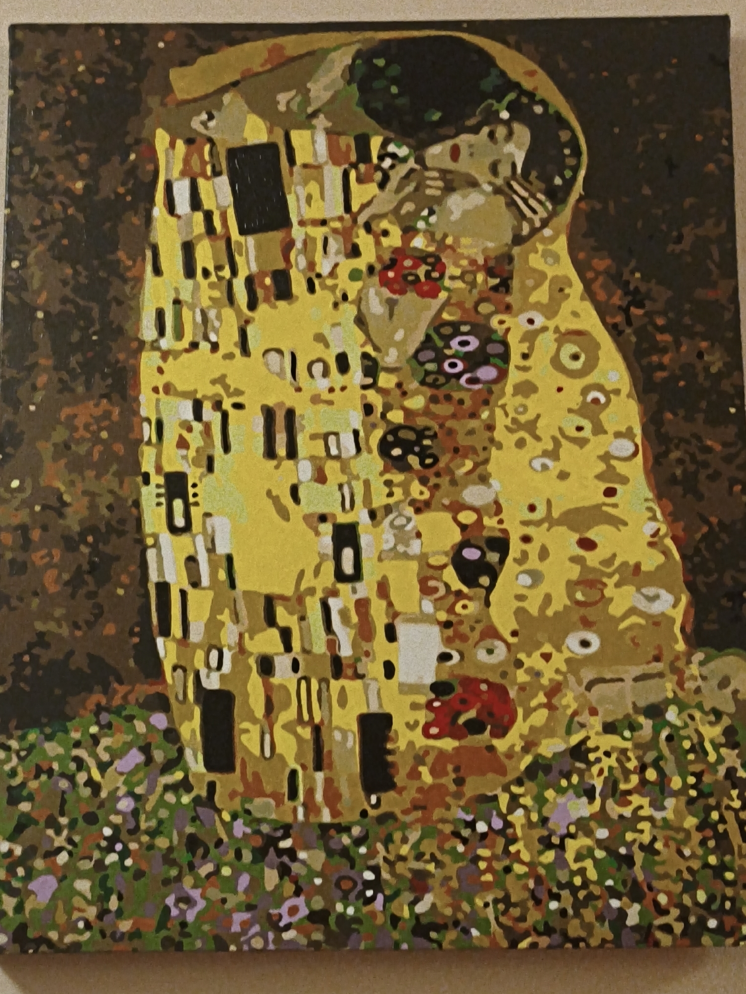 The image is a reproduction of Gustav Klimt's painting "The Kiss". It depicts a couple embracing in a golden robe, set against a patterned background. The painting is rendered in a style that appears to be paint-by-numbers, with distinct areas of color and simplified shapes. The background features abstract, organic shapes and speckles of brown, black, and gold. The foreground is a textured expanse of green, purple, and yellow, suggesting a floral meadow. The couple's forms are largely obscured by their elaborate, patterned robes, which are decorated with squares, circles, and spirals. The man's head is bowed, kissing the woman's cheek, while her eyes are closed. This particular reproduction is not located in Steblivka, Ukraine; it is a painting.