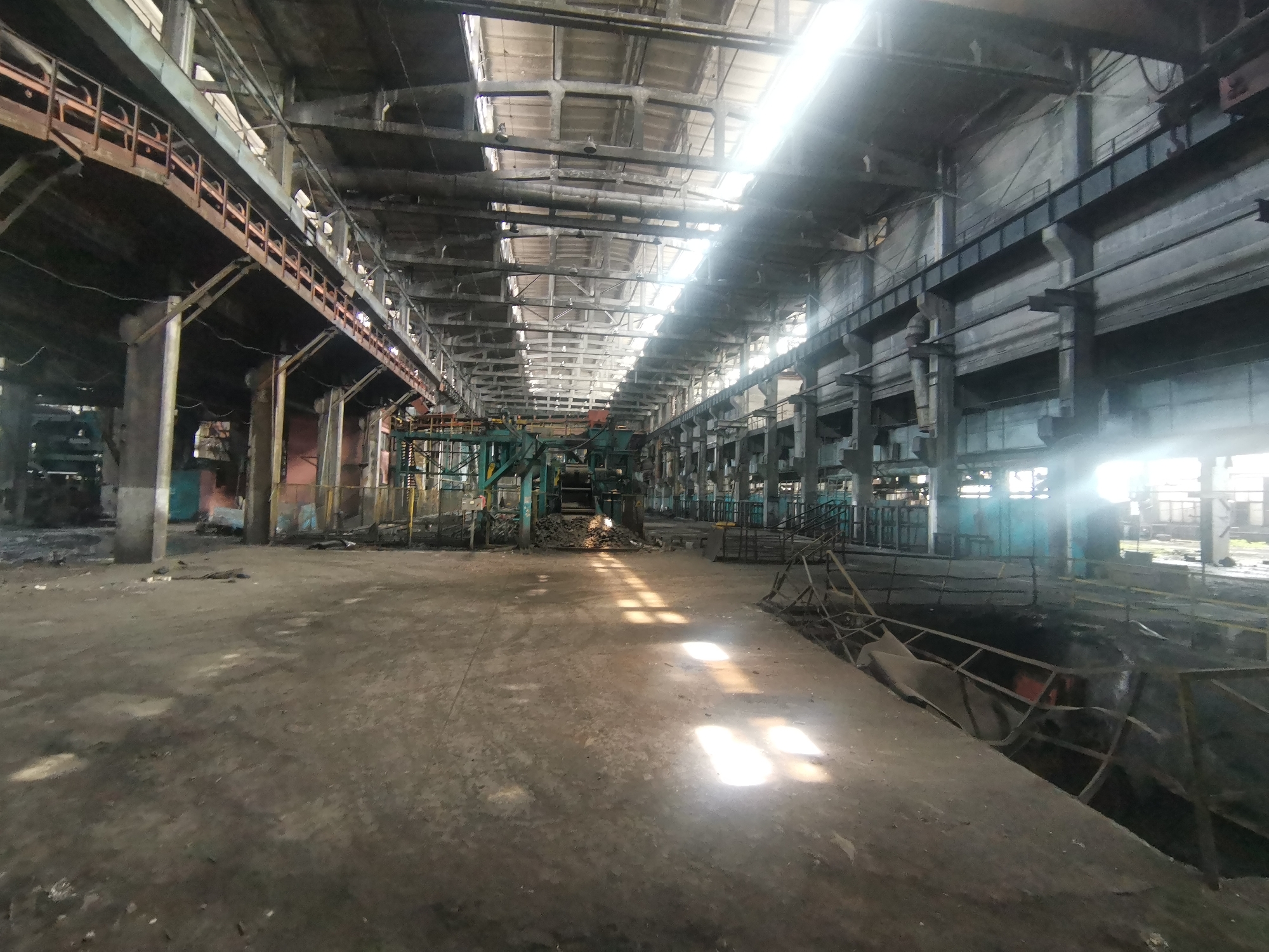 The image depicts the interior of a large, abandoned industrial building. Light streams in through the roof, illuminating a vast concrete floor covered in debris. A rusting metal structure stands in the middle ground, suggesting machinery remains. The building's architecture features high ceilings, support pillars, and metal beams. The overall scene suggests a state of disuse and decay. There is no evidence of people, alive or deceased. There is no information available in this image to connect this building to Zhivkovo, Bulgaria.