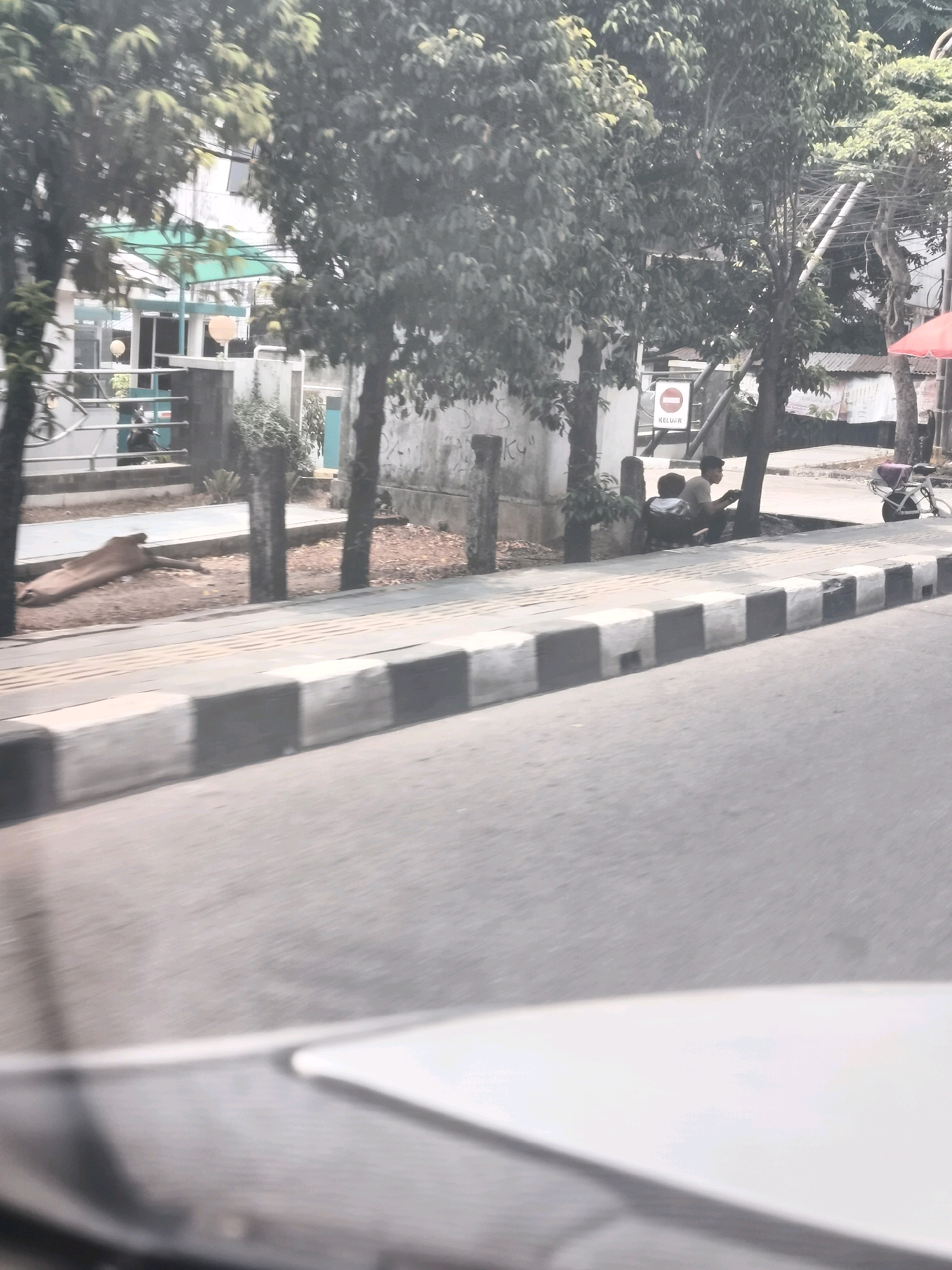 The image shows a street scene in Palembang, Indonesia. In the foreground, there is a paved road with a sidewalk to the left. The sidewalk has a distinctive pattern of alternating black and white rectangular blocks. Behind the sidewalk, there are several trees casting shadows. In the middle ground, there is a low wall and a building with a green awning. A few people are sitting by a tree next to a sign that says "KELUAR", which means "EXIT" in Indonesian. A bicycle is parked near the people. In the background, there are more buildings and trees. The overall impression is a sunny day in a city.
