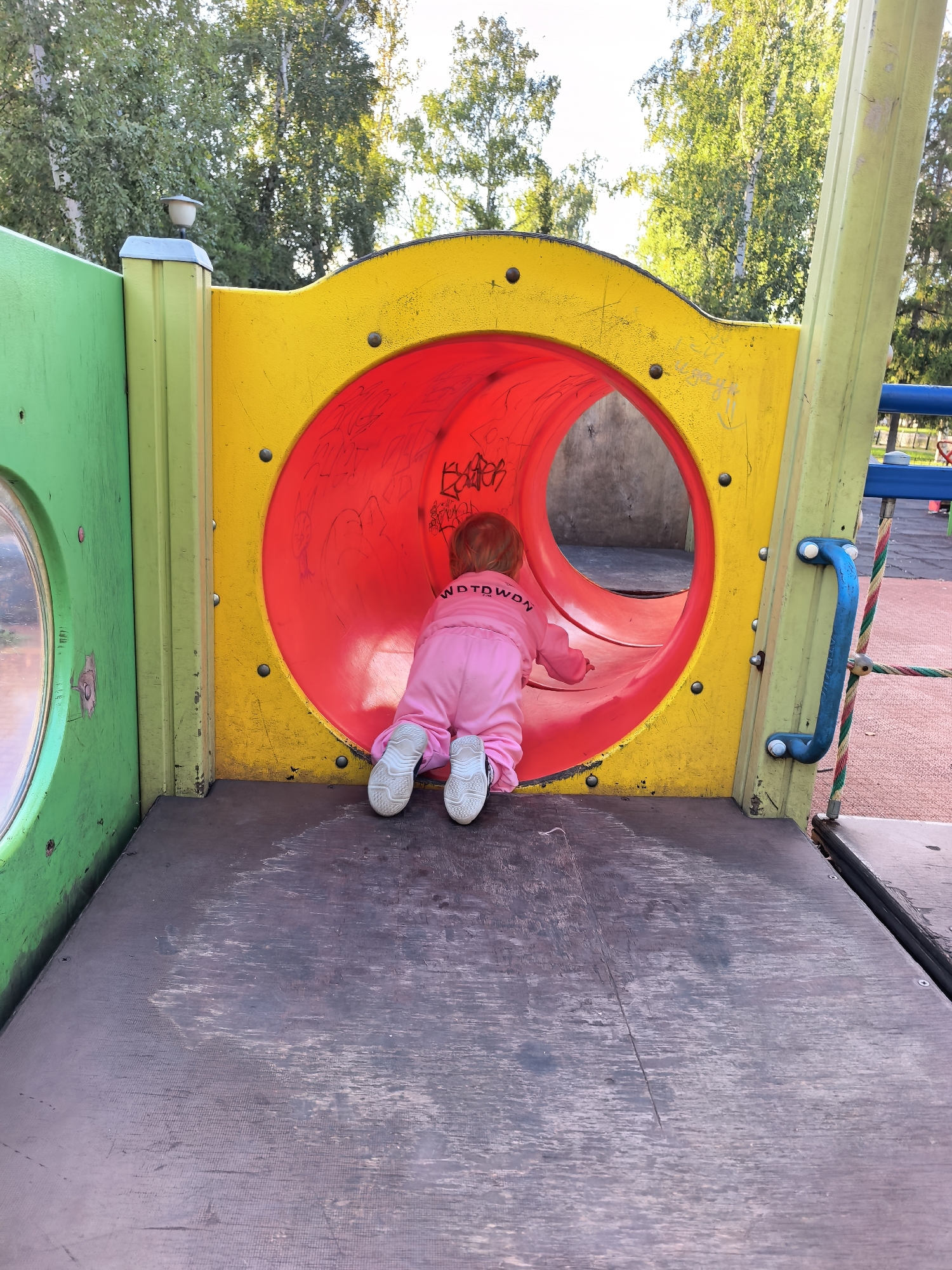 An overhead view captures a toddler wearing a pink jumpsuit and white sneakers on a wooden platform of a playground. The toddler is crawling into a red, circular tube tunnel of a yellow and green playground structure. The tunnel is adorned with graffiti. The playground is surrounded by trees with green foliage, suggesting an outdoor setting. The ground surface is a mix of wooden planks and what appears to be red rubber safety surfacing. The image lacks any identifiable landmarks or context that would place it specifically in Sterlitamak, Russia.