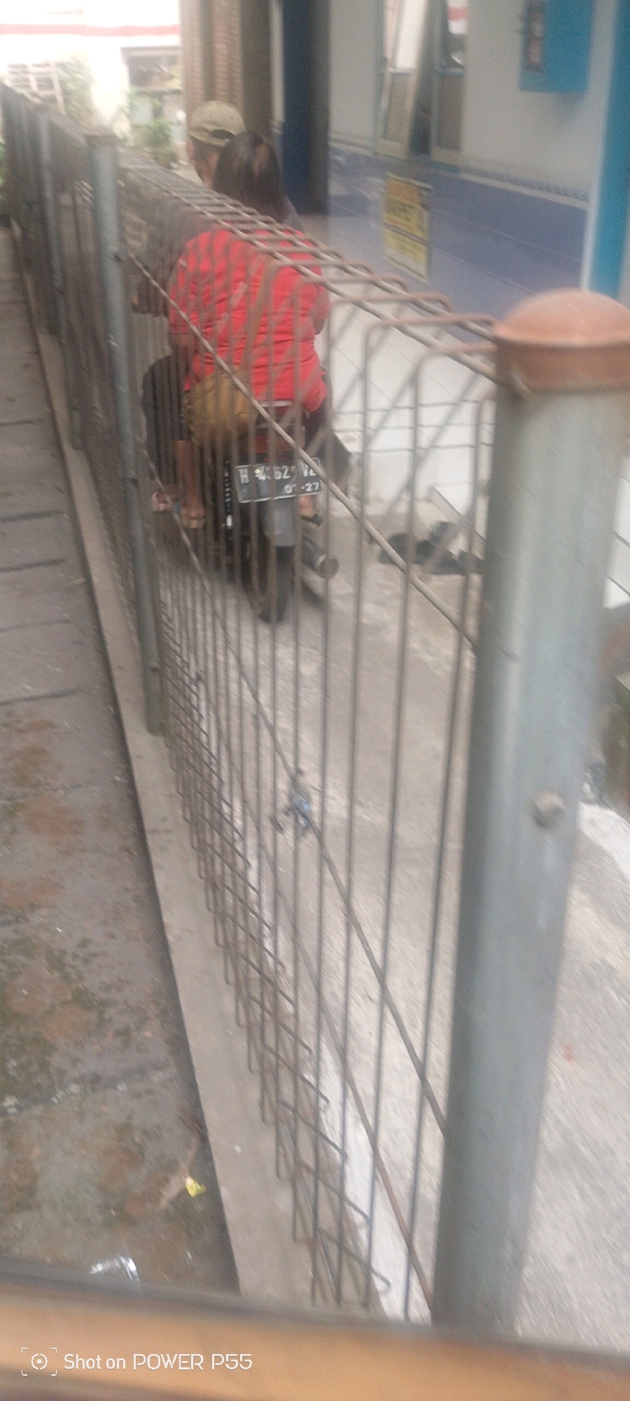 The image, taken in an urban setting, shows two individuals on a motorcycle. The person in the foreground wears a red shirt, and the person behind them, partially visible, wears a cap and a light-colored shirt. The motorcycle's license plate reads "H 4362 VZ". A metal fence, appearing to be wire mesh, occupies the foreground, obscuring parts of the scene. The ground is paved with concrete. In the background, a building with blue and white tiled walls is visible. The overall lighting suggests it is daytime.