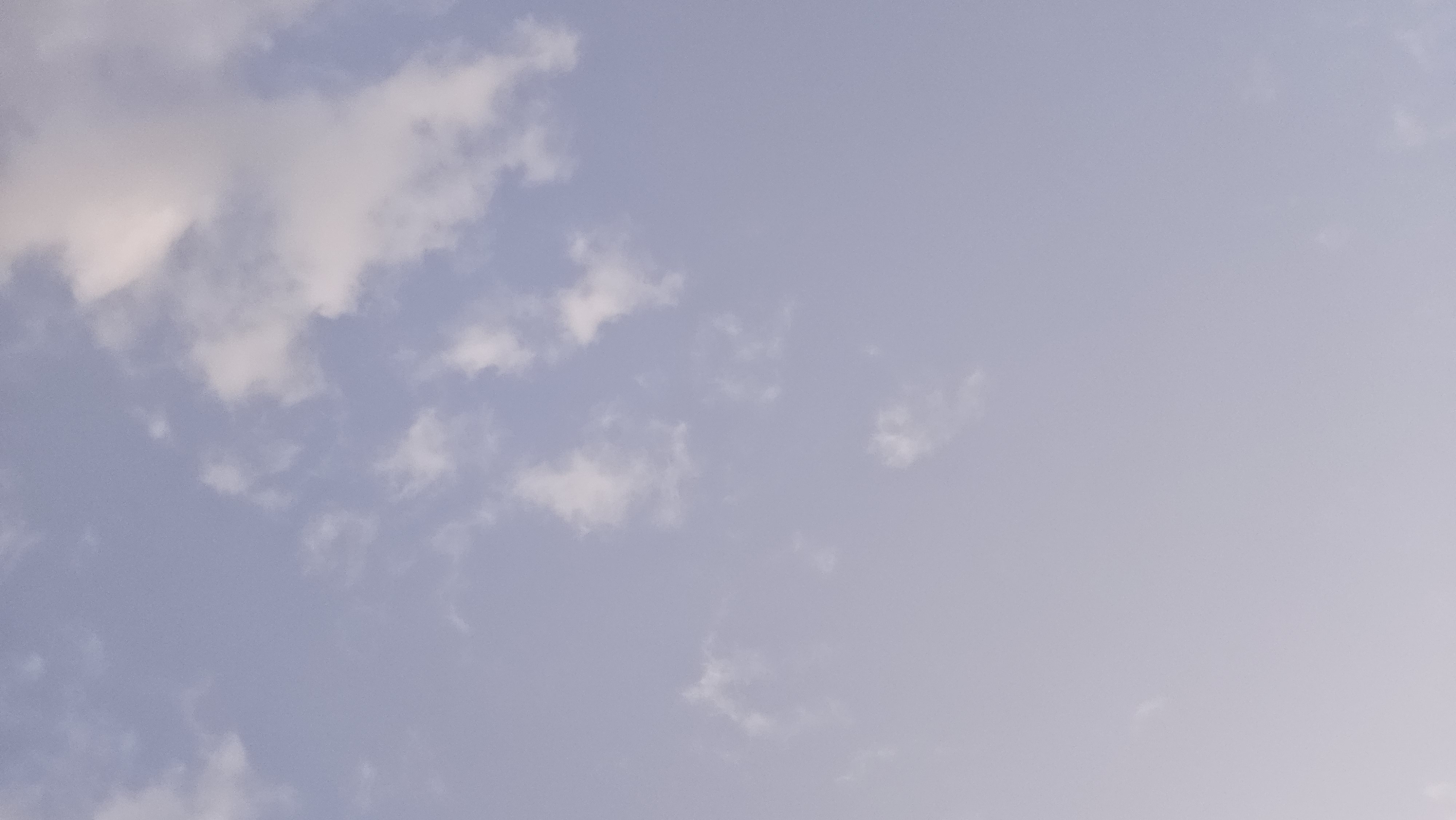 The image is a vertical, low-angle shot of a pale blue sky with scattered white, wispy clouds. The clouds are small and irregularly shaped, with some appearing brighter and more defined than others. The lighting suggests it is daytime, possibly late afternoon or early morning, with the sun diffused by the clouds. There are no discernible objects or structures visible in the frame, and the scene appears to be an open sky. The location is described as Unknown, Unknown.
