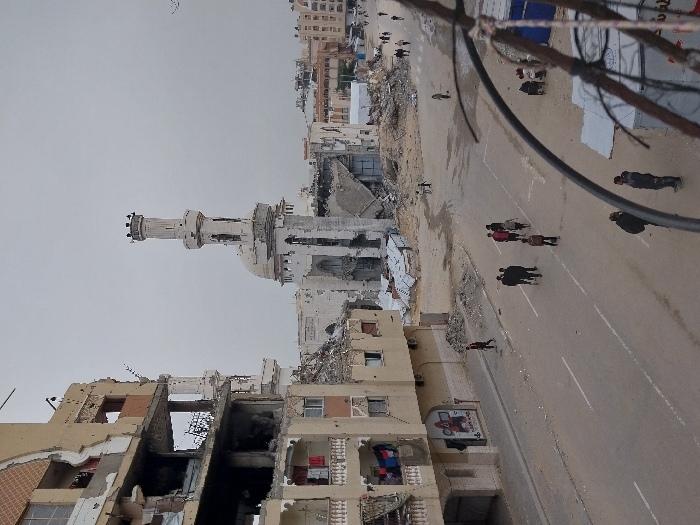 The image portrays a devastated urban landscape in Khan Yunis, Palestinian Territory, under an overcast sky. In the center, a severely damaged mosque features a tall, slender minaret, largely intact at its apex but extensively compromised along its shaft. The mosque's dome and main structure are partially collapsed, reduced to large piles of rubble and debris.
To the left, a multi-story building exhibits extensive structural damage across multiple floors, with significant portions of its exterior walls destroyed, exposing interior rooms. Laundry is visible hanging on a balcony on one of the lower, partially intact floors.
The foreground shows a wide, paved road with white lane markings. Multiple individuals are present on the street; a group of approximately 6-8 people walks centrally, while other individuals walk or stand closer to the sides and in the distance. Further into the background, more people are seen navigating through paths cleared amidst widespread rubble and the remnants of destroyed buildings. Debris, including concrete chunks and twisted metal sheets, is scattered across the ground, particularly around the damaged structures. In the extreme bottom right, parts of a white and red temporary structure are visible, partially obscured by dark cables and wires.