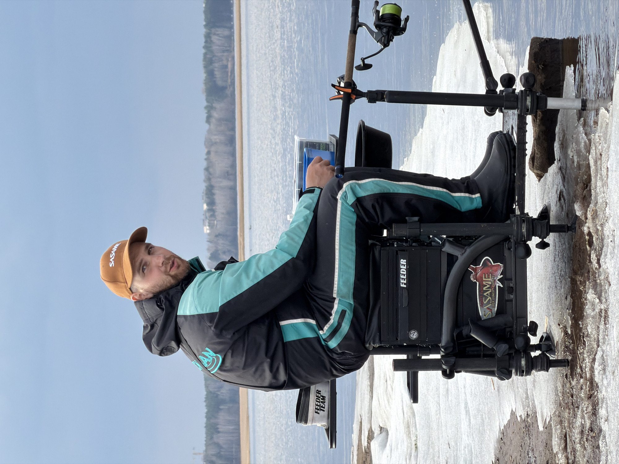 A man is ice fishing on the Kama River in Chaykovsky, Russia. He is seated in a black fishing chair with the word "Feeder" on its side, wearing a black jacket with light blue accents and a brown "Sasame" baseball cap. He is holding a blue container. In front of him are two fishing rods, one with a green fishing reel. The ground is covered in ice and snow. In the background, the river is visible, along with the opposite shore. The sky is light blue.