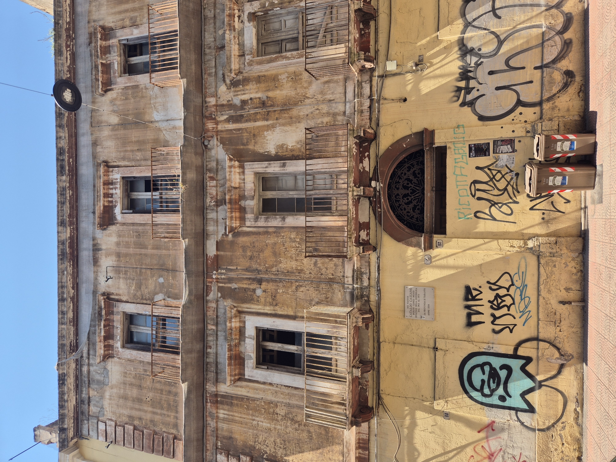 The image depicts a weathered, multi-story building in Taranto, Italy, characterized by its peeling facade, rust-stained balconies, and multiple windows, some boarded up or showing signs of disrepair. The lower portion of the building is painted a yellow hue and extensively covered with graffiti. Visible graffiti includes a blue ghost-like figure with large eyes and buttocks, stylized text, and larger, more abstract tags. Two brown recycling bins are situated in front of the building. A plaque, likely a historical marker, is affixed to the yellow wall. The scene is set on a sunny day, with shadows cast across the lower section of the building. The overall impression is one of urban decay and neglect, juxtaposed with the vibrant, albeit illicit, street art.