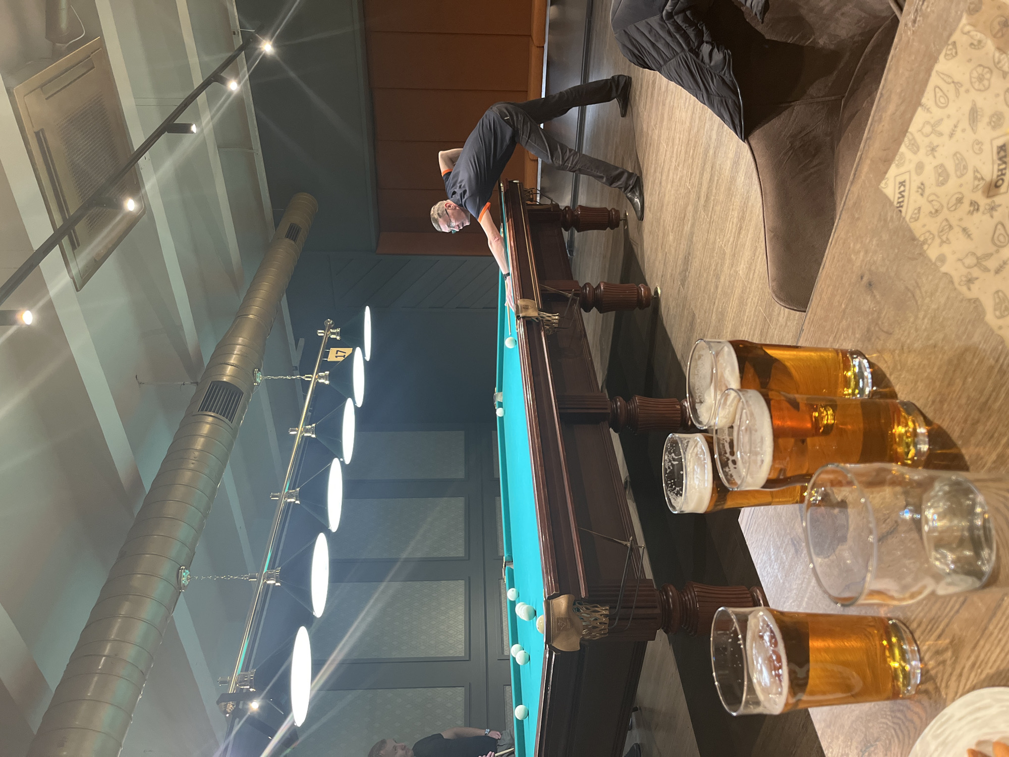 The image captures a scene in what appears to be a bar or pub with a billiards table. A man is bent over the pool table, aiming or taking a shot. The billiard table is set up with multiple balls. Several glasses filled with a golden-colored liquid, likely beer, are situated on a table nearby. The bar has long, silver light fixtures hanging above. The exact location of this image is in Izhevsk, Russia.