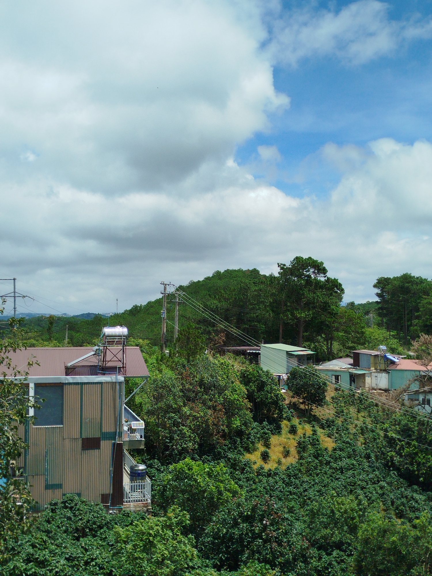 The image depicts a rural or semi-rural hillside settlement in Đa Thọ, Vietnam, characterized by lush green vegetation and a scattering of buildings. The foreground is dominated by dense trees and bushes, suggesting a natural environment. In the middle ground, several structures are visible. On the left, a multi-story building with a corrugated metal exterior is partially obscured by foliage. To its right, and slightly uphill, a brown-roofed house features a prominent silver water tank on its roof, supported by a metal frame. Further to the right and uphill, a series of smaller buildings with light green and blue-colored walls and roofs are nestled into the slope. These buildings appear to be residences, with some exhibiting signs of everyday use, such as laundry hanging outside. The landscape rises behind the buildings, covered in more dense green trees, likely a forest or plantation. Overhead, a vast sky with scattered clouds, some bright white and others darker, suggests a partly cloudy day. Power lines are visible crisscrossing the scene, connecting utility poles that are integrated into the landscape. The overall impression is one of a community living in harmony with a verdant, hilly environment.