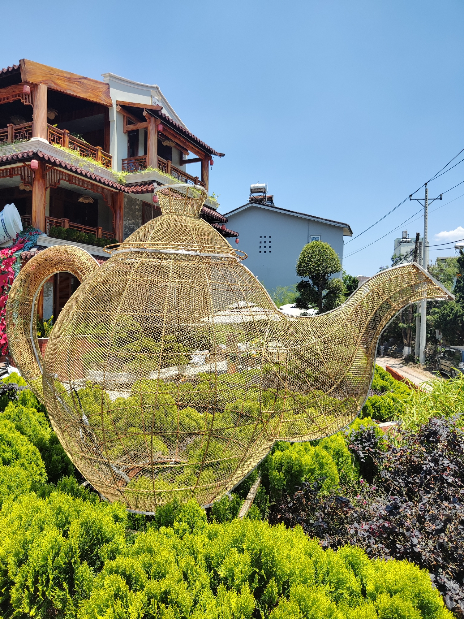The image showcases a large, golden, wireframe sculpture of a teapot in Da Lat, Vietnam. The teapot is set amidst a lush garden of small, green, spherical bushes and some dark purple foliage. The wireframe structure is intricate, mimicking the shape of a classic teapot with a rounded body, a curved handle, and a spout. Inside the wireframe, glimpses of the surrounding greenery can be seen.
Behind the teapot, there is a building featuring traditional Vietnamese architectural elements, including wooden beams, ornate railings, and curved eaves. A portion of a white water tank is visible on top of the roof and a house with gray walls and a solar water heater on the roof. Power lines stretch across the sky. The sky itself is bright blue, indicating clear weather.