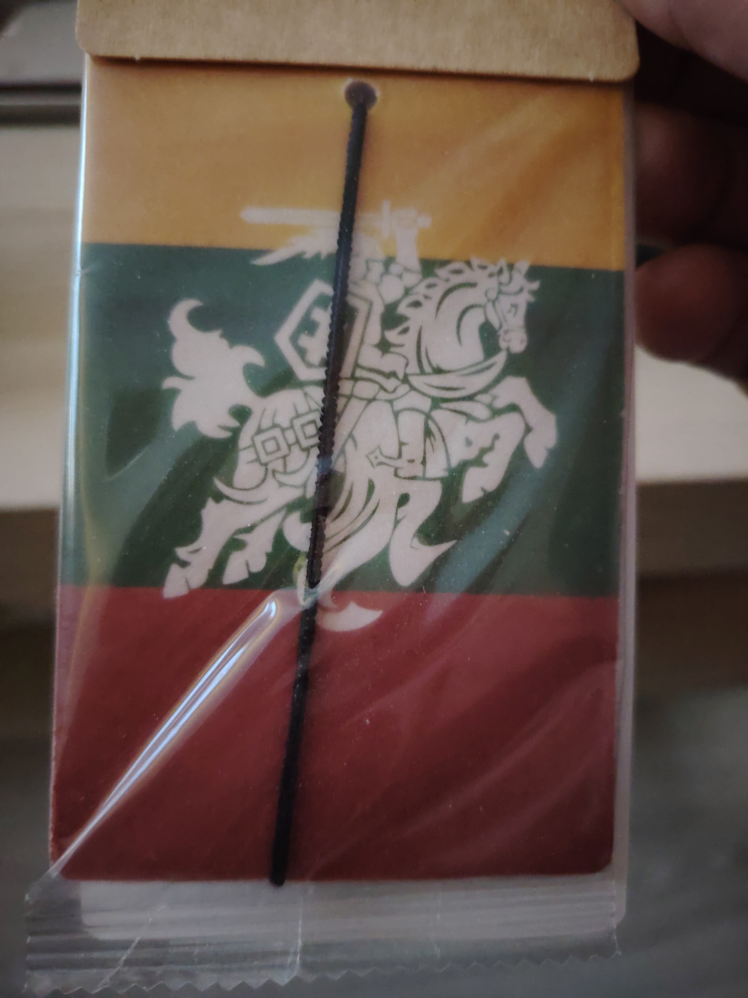 This is a close-up, eye-level, indoor shot of a small, rectangular item, likely a bookmark or a small flag, encased in clear plastic. The item features the flag of Lithuania, with horizontal stripes of yellow at the top, green in the middle, and red at the bottom. A white depiction of the Lithuanian coat of arms, known as the Vytis, is prominently displayed across the green and red sections. The Vytis depicts a knight on horseback, armed with a sword and shield, in pursuit of an enemy. A thin black string or cord is attached to the item, running vertically down the center. The background is blurred and indistinct, suggesting an indoor setting, possibly a retail environment or a personal collection. The lighting is moderate, with some glare on the plastic casing. No specific location in Huntingdonshire, United Kingdom can be determined from the image.