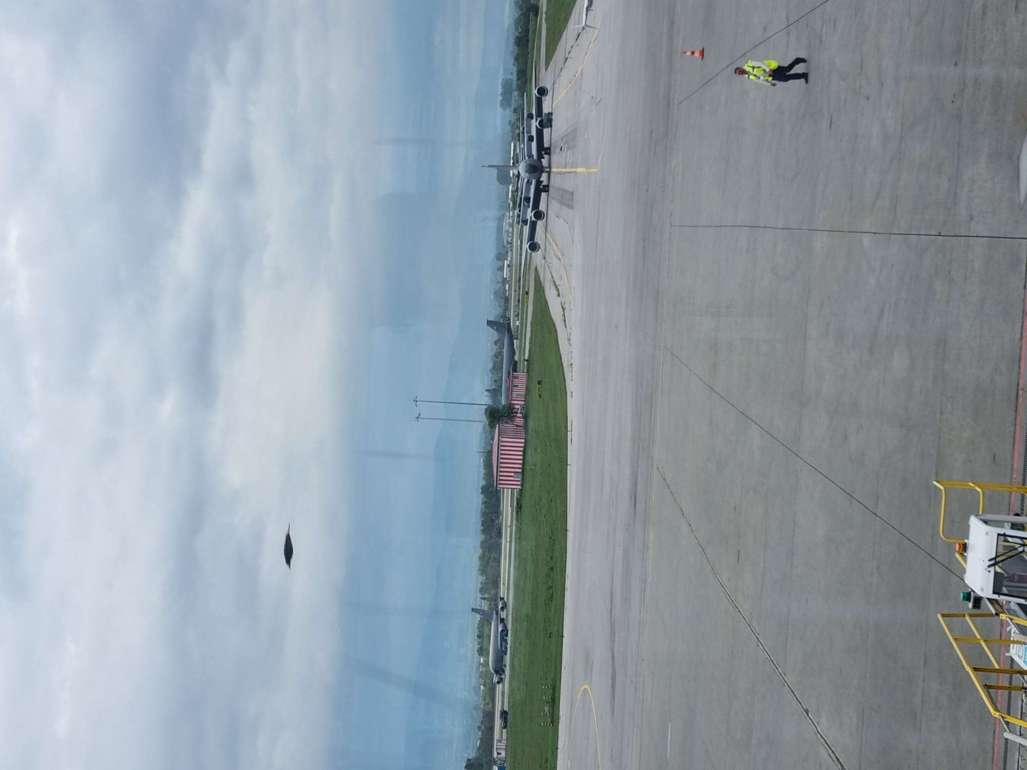 The image depicts an airport scene, likely from a terminal window. The location is Sofia, Bulgaria, based on the presence of aircraft resembling the KC-135 Stratotanker, often associated with military facilities in Eastern Europe.
Several aircraft are visible on the tarmac, lined up. A ground crew member wearing a high-visibility vest is walking on the concrete apron. A small cone is nearby. A section of an airport boarding bridge is visible on the right. The sky is overcast with visible clouds. The environment is an active airport area.