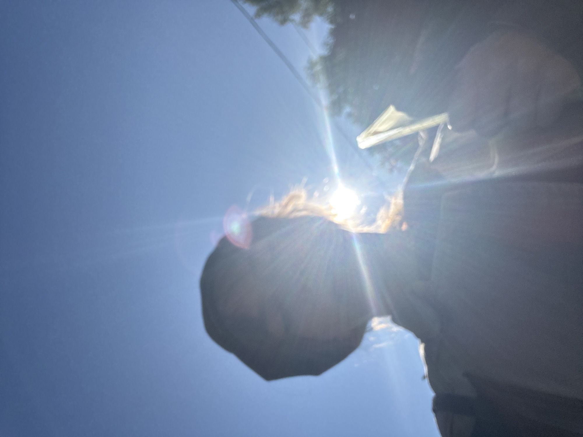 A person, silhouetted against a bright blue sky, holds a folded piece of paper or thin material in their right hand. The sun is positioned behind the person's head, creating a strong lens flare that obscures facial features and casts rays of light across the image. The person appears to be wearing a dark hat and a light-colored shirt. The background consists of the sky and some out-of-focus trees and power lines, suggesting an outdoor urban or suburban environment. The location is identified as Novyy, Russia.