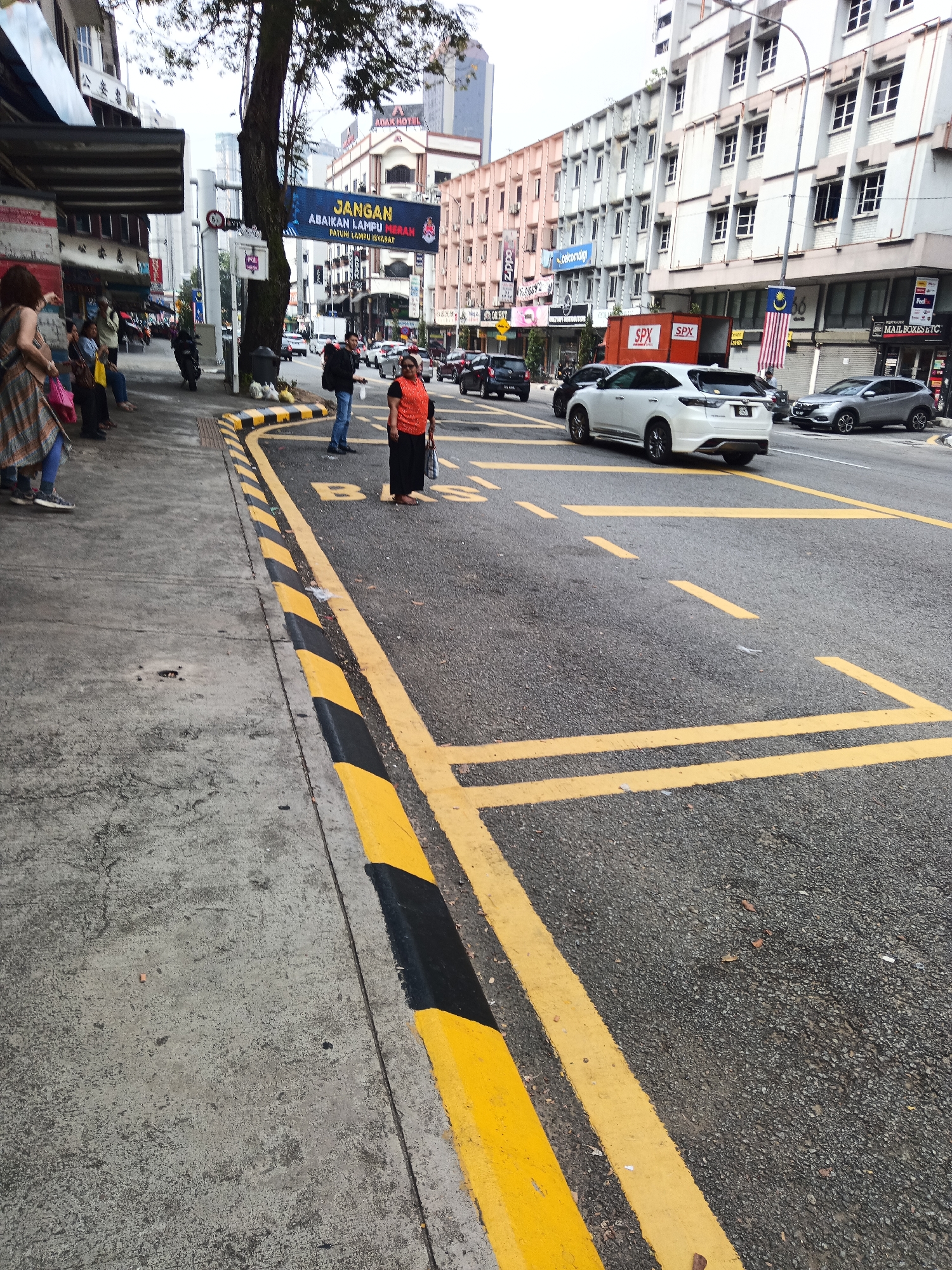 The image shows a street scene in Kuala Lumpur, Malaysia, with a focus on a bus stop area. In the foreground, a yellow and black striped curb runs along the edge of the sidewalk. The sidewalk itself is made of concrete and appears worn. Yellow lane markings on the asphalt road indicate a bus stop zone.
Several people are visible at the bus stop. On the left side of the sidewalk, a woman in a patterned dress and blue jeans is standing with her back to the camera, holding a bag. Next to her are other individuals, some of whom are also standing or sitting.
In the road, a woman in an orange top and black skirt is standing near the bus stop markings, holding a bag. A man in a blue shirt and jeans is standing next to her. A white SUV is driving on the road, and other vehicles are present in the background.
The background shows a row of multi-story buildings with various signs and awnings, typical of a commercial street in Kuala Lumpur. A large banner with the text "JANGAN ABAIKAN LAMPU MERAH PATUHI LAMPU ISYARAT" (Do not ignore red lights, obey traffic signals) is prominently displayed on a building. The sky is overcast. The overall environment suggests a busy urban setting.