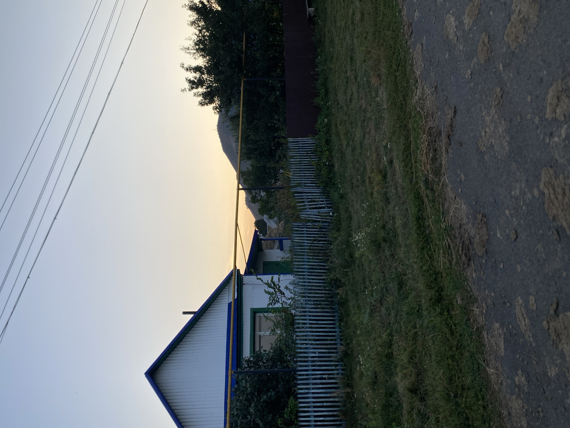 The image depicts a rural scene in Maloarslangulovo, Russia. The main subjects include a white house with blue trim and a green-trimmed window, a blue and white striped fence, a dark brown fence, and a gas pipe. The setting is outdoors, with a grassy area to the right of the fences and a dark, textured road or path in the foreground. In the background, trees and a distant hill are visible, with power lines stretching across the sky. The sky is a pale, hazy blue, suggesting either early morning or late afternoon. The lighting indicates it is likely dusk or dawn. There are no people or animals visible in the image, nor is there any text. The overall mood is quiet and somewhat serene, characteristic of a rural landscape at the end or beginning of the day.
