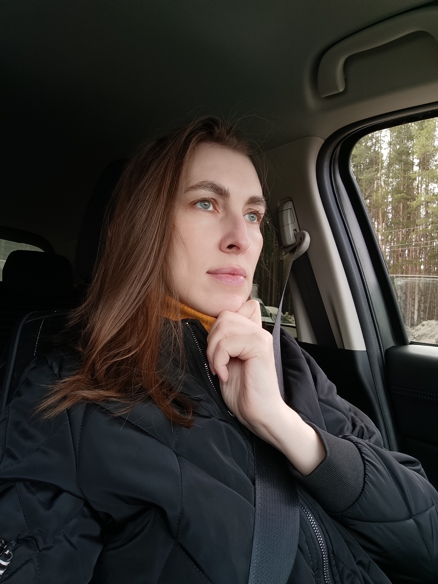A woman with auburn hair, wearing a yellow turtleneck sweater and a black quilted jacket with a seatbelt across her chest, is seated in a car. She is resting her chin on her hand and looking out the window. Outside the car window, trees and a snow-covered ground are visible. The location is presumably near a wooded area. The photograph may have been taken in or around Votkinsk, Russia, but there is no specific context to confirm this.
