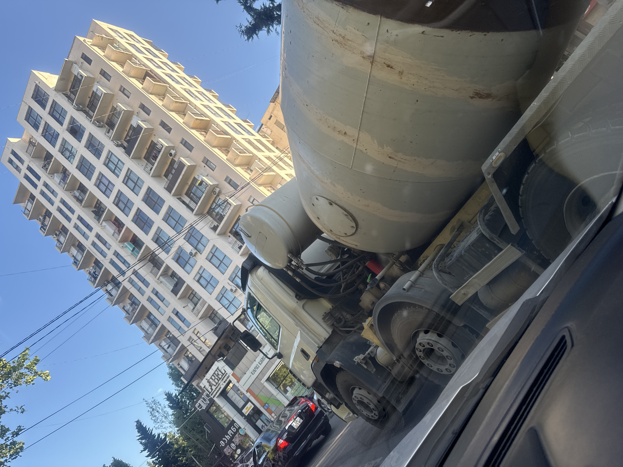 The image depicts a street scene in Tbilisi, Georgia, on a clear, sunny day, likely late morning or early afternoon given the bright light and distinct shadows. The primary subjects are a large concrete mixer truck and a modern, multi-story apartment building.
The apartment building, prominently featured on the left side of the frame, is light-colored with numerous balconies and windows. Its architecture suggests a contemporary urban setting. In front of the building, a concrete mixer truck dominates the right side of the image. The truck is positioned such that its large mixing drum is in the foreground, angled upwards. The lower portion of the truck's chassis, wheels, and cab are also visible.
The scene appears to be a typical urban street with other vehicles, including a dark-colored car in the middle ground, suggesting ongoing traffic. There are also trees and overhead power lines, common elements in cityscapes. Reflections on the truck and potentially on a vehicle from which the photo is taken add a layer of depth and visual interest.
Visible text on signage in the middle ground includes "LABEL" in English, and some text in Georgian script, indicating commercial establishments. The overall impression is that of a bustling urban environment captured from a unique perspective, possibly from another vehicle. No people are clearly discernible. The atmosphere is bright and clear, indicative of good weather.