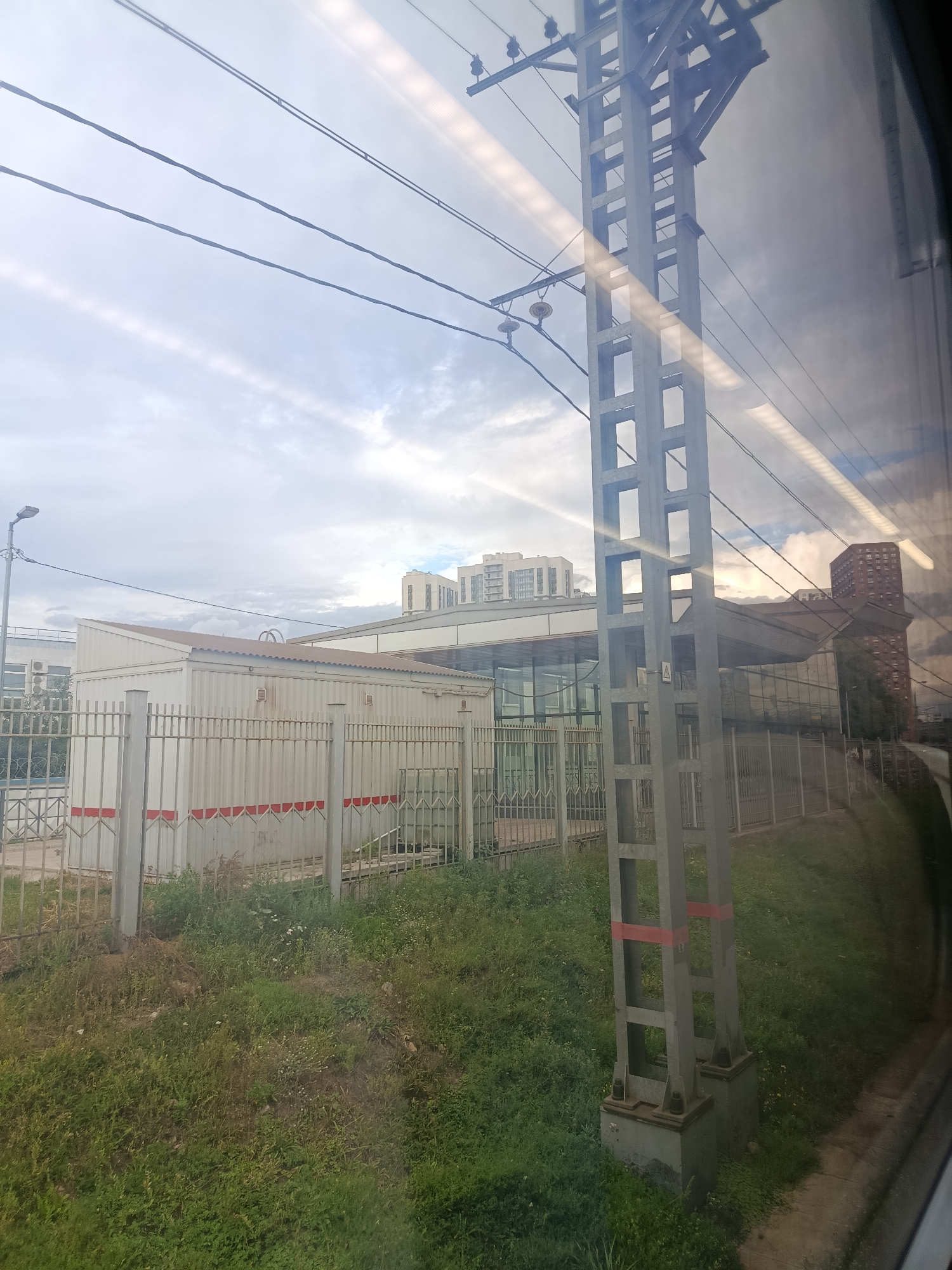 The image depicts a view from a train window, likely in Moscow, Russia, due to the presence of typical Russian urban infrastructure. In the foreground, a tall, grey metal electricity pylon with red banding stands prominently on the right side, a common sight along railway lines. The pylon supports multiple overhead power lines that stretch across the sky. To the left of the pylon, a long, white, corrugated metal building with a slightly sloped roof is visible behind a metal fence. The building appears to be some kind of utility or maintenance structure. Beyond this building, a modern glass-fronted structure with a curved roof and several tall residential buildings can be seen in the distance. The sky is overcast with streaks of light suggesting the sun is attempting to break through the clouds. The ground in front of the fence is covered in green grass and some weeds. The reflection of the interior of the train is visible on the train window, particularly on the right side of the image, distorting some of the view.