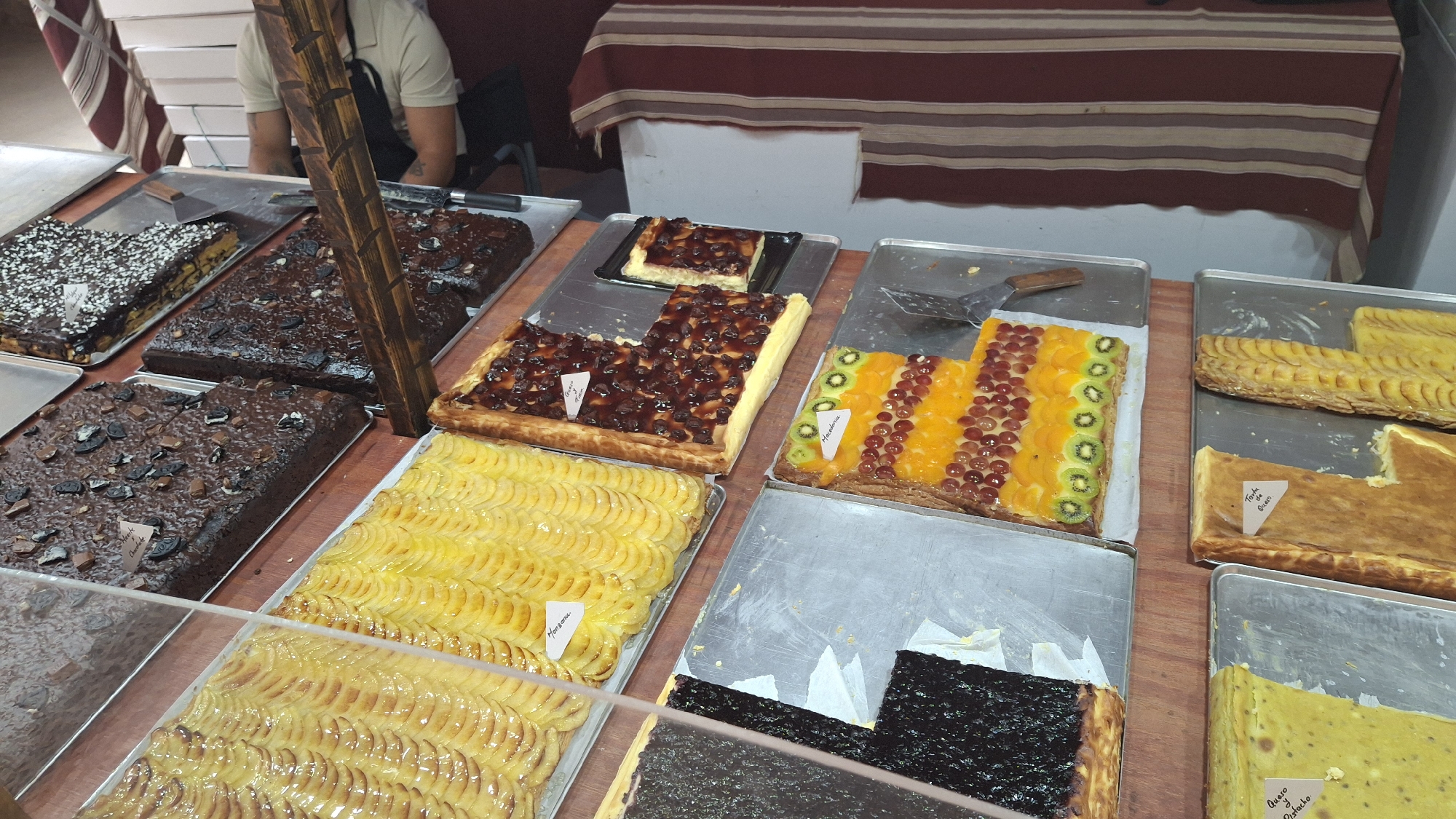 In a bakery in La Fresneda, Spain, a display of assorted pastries is presented. The pastries are arranged on metal trays and include what appears to be chocolate cakes, fruit tarts with grapes and kiwi, and thinly sliced apple tarts. A wooden display stand is positioned between some of the trays. In the background, a person wearing a light-colored shirt and a dark apron is partially visible, suggesting they are a worker in the establishment. The overall scene is one of a retail environment offering baked goods for sale.