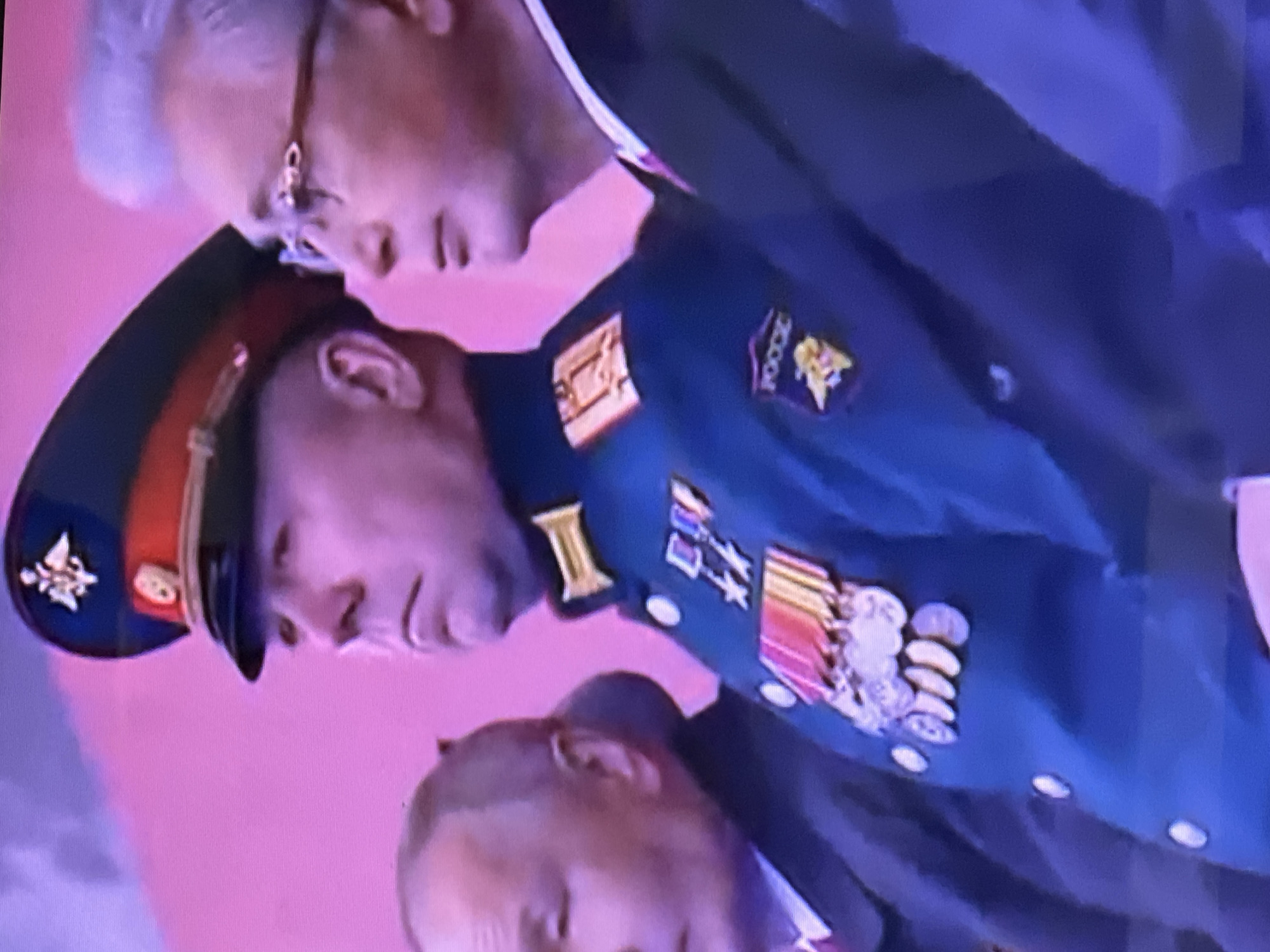 The image shows three individuals. The central figure is a man in a dark military uniform with numerous medals and insignia, including a badge with "Россия" (Russia) written on it. Above him is an older man in a suit and glasses. Below, there is a partial view of another man's head. The background is a blurred, light pink color. The specific location in Chaikovsky, Russia, is not discernible from the image.