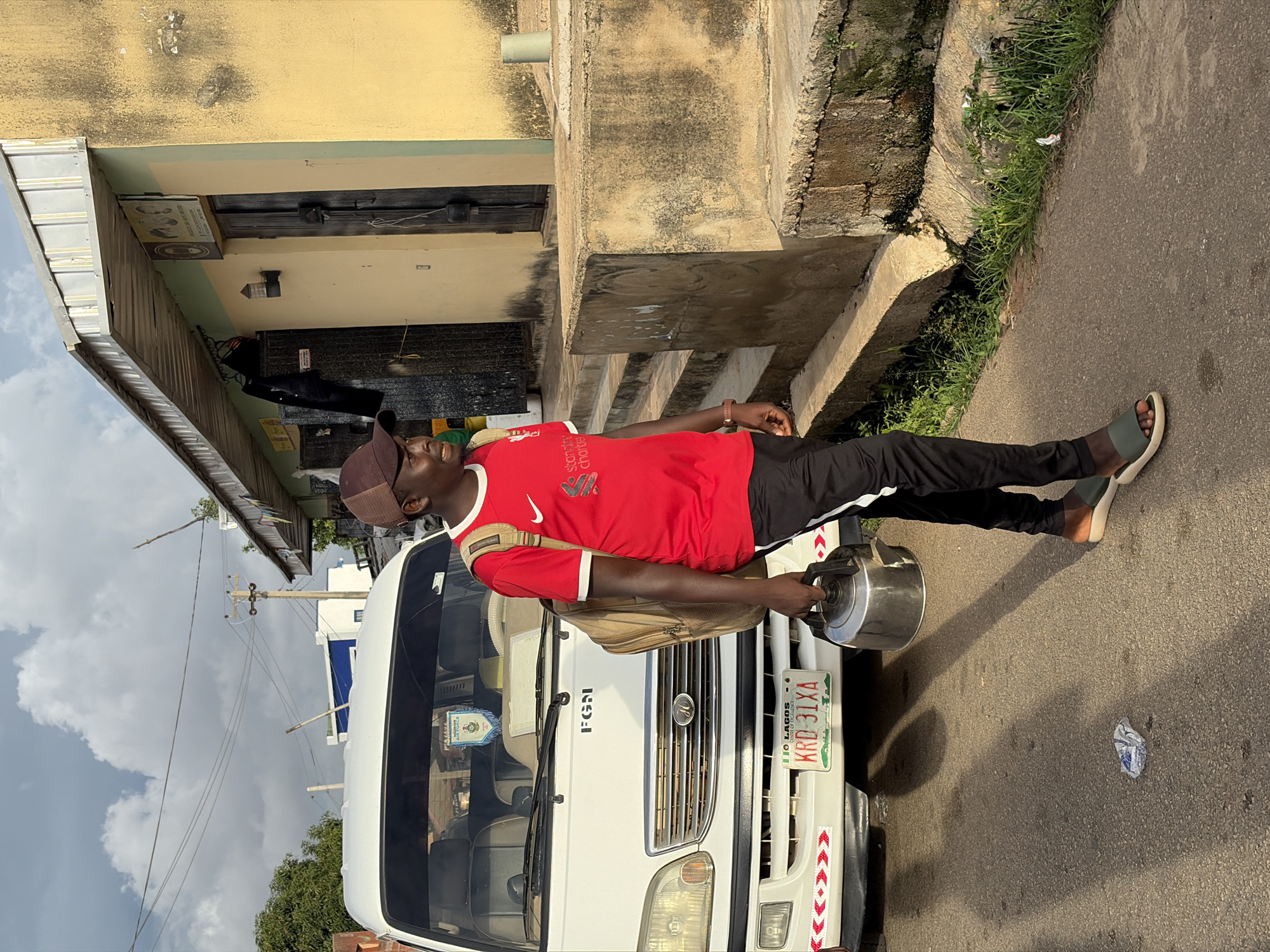 This image depicts a street scene in what is described as Jos, Nigeria, under a cloudy sky. A young, dark-complexioned male walks from left to right on a paved asphalt road, carrying a tan backpack and a silver metal kettle in his right hand. He wears a red short-sleeved t-shirt with a white collar, a white Nike swoosh, and "Standard Chartered" branding, dark track pants, a brown baseball cap, and olive-green slide sandals. His shadow is cast on the road ahead of him.
To the left of the male, a white utility van or minibus is parked, facing the viewer. The vehicle has "FGN" printed on its front grille, an "H" shaped emblem, and a Nigerian license plate from Lagos reading "KRD 31XA." A blue and white pennant displaying "NIGERIAN AIRFORCE" is visible inside the windshield. The front bumper is adorned with red and white chevron-patterned tape.
On the right side of the frame, a building with a light beige/yellow facade and a light green upper section is present, topped by a corrugated metal roof. A wooden door with an exterior light fixture is visible, above which hangs a sign with images and text. A raised, weathered concrete platform with steps borders the road, separating it from the building. To the left of the main door, a dark wooden gate or door is partially visible. The immediate roadside and concrete drainage channel exhibit patches of grass and weeds, along with a crumpled white wrapper on the asphalt. Overhead power lines are visible against the cloudy sky, and a utility pole is present in the mid-ground.