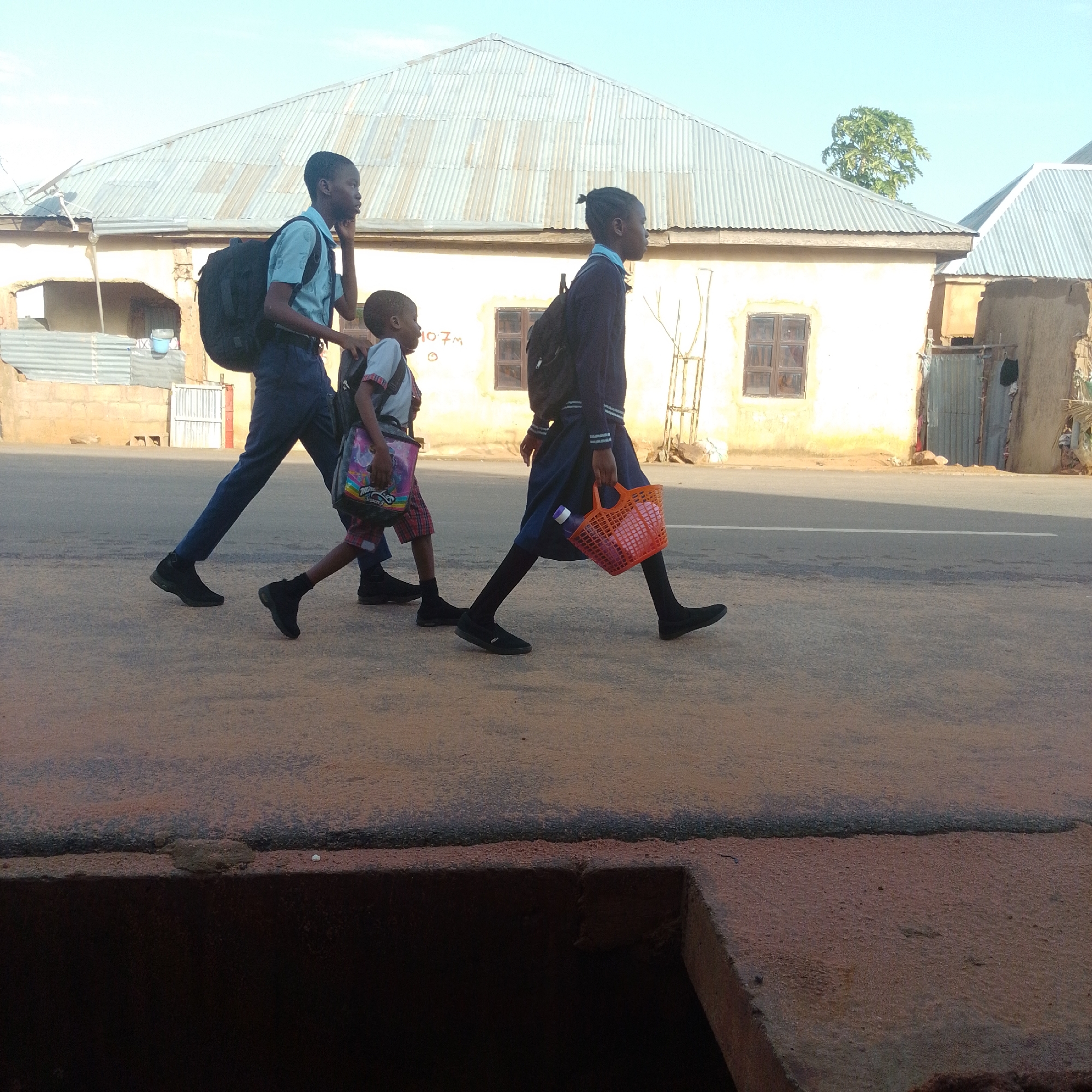 In Tempure, Nigeria, three young school children, two boys and a girl, walk on a dirt road alongside a paved street. The boy in the lead, wearing a light blue shirt and dark pants, carries a black backpack. The second child, a boy in a checkered skirt and light blue and red shirt, also has a backpack. The third child, a girl in a dark blue uniform and black tights, carries an orange basket filled with items, including a purple bottle. The children are walking from right to left in the frame. Behind them, a light-colored building with a corrugated metal roof is visible, with several windows. The sky is clear and bright blue. The foreground shows a dark, textured surface, possibly a drainage channel, with a lighter brown, dusty area above it.