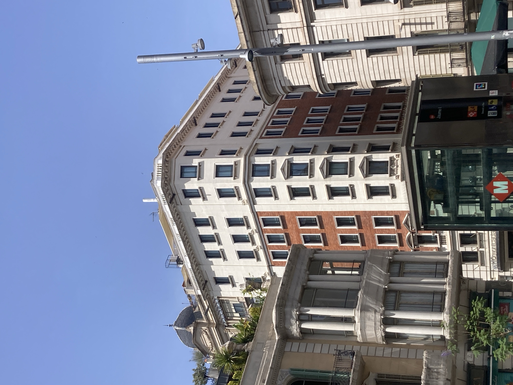 The image shows a street scene in Barcelona, Spain, dominated by the architecture of several multi-story buildings. The buildings feature a mix of classical and modern design elements, with ornate balconies, large windows, and brick and stucco facades. In the foreground, there is a glass structure that appears to be an entrance to a metro station, indicated by a prominent "M" logo. The sky above is clear and blue. The buildings are several stories high and exhibit a range of architectural styles, from the rounded facade of the building on the right to the more angular structure in the center. The building on the far left has a dome structure. There are no people clearly visible in the image.