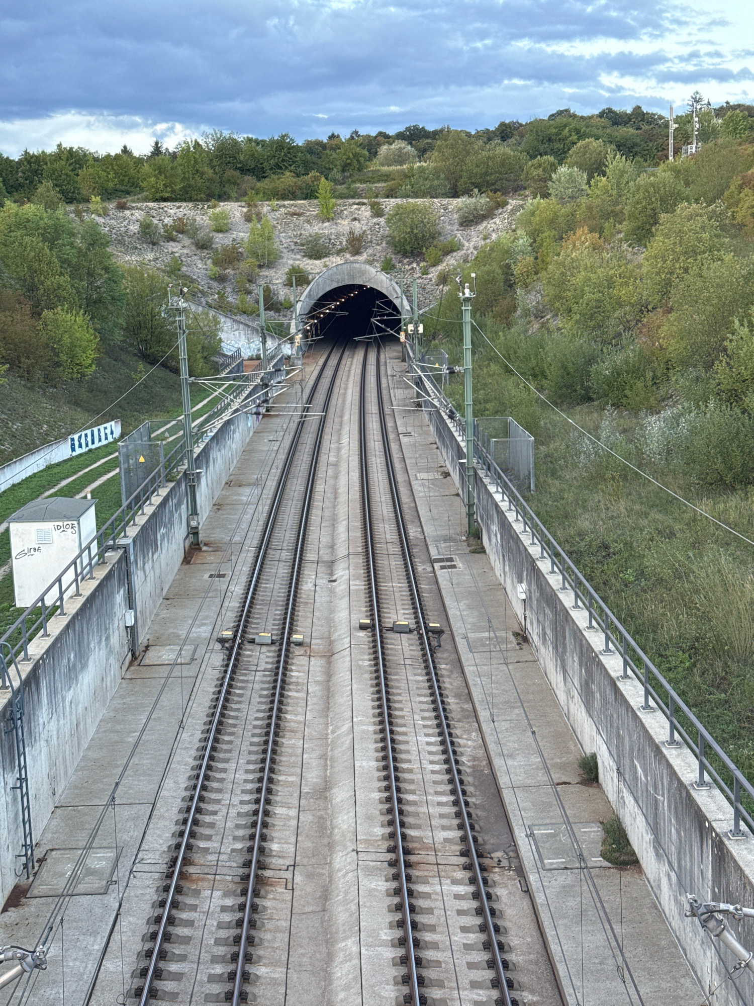 The image shows a view of a double-track railway line leading into a tunnel. The railway tracks are made of steel rails and concrete sleepers, and they are positioned on a concrete bed. There are overhead power lines for the electric trains. On either side of the tracks are concrete walls with railings, separating the railway from the surrounding environment. The area around the railway is covered with vegetation, including grass and trees. In the background, there is a hill with more trees and a cloudy sky. The tunnel entrance is a concrete archway, and lights are visible inside the tunnel. There is a small white building with graffiti on the left side of the image. This scene is located in Wettstetten, Germany. There are no people visible in the image.