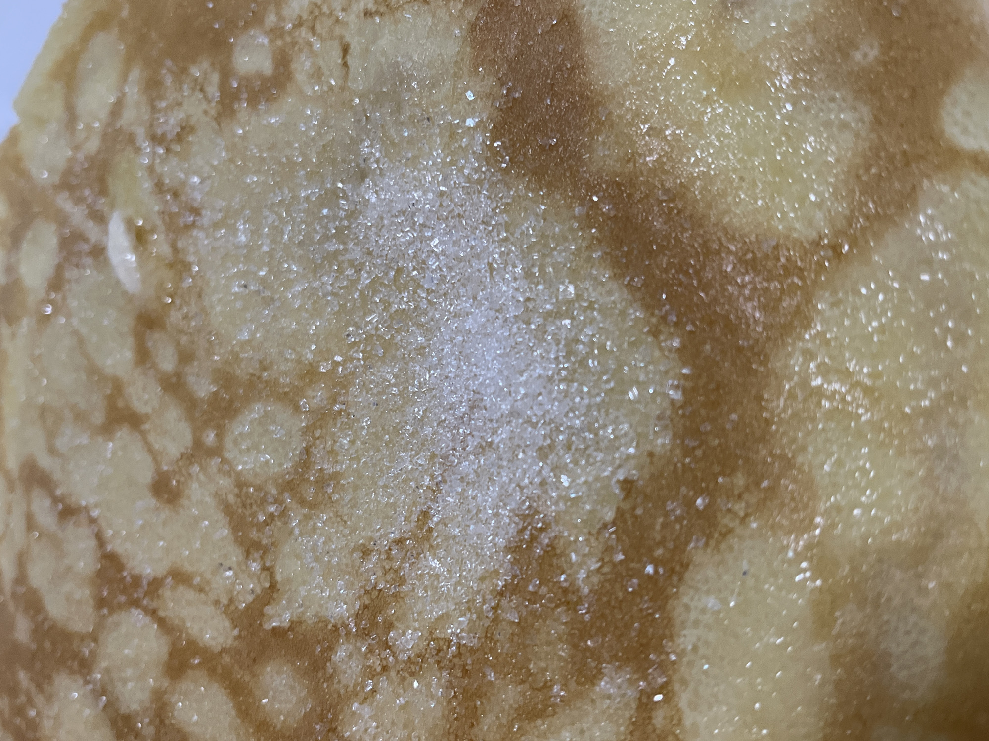 A close-up, top-down shot of a pancake covered in granulated sugar. The pancake has a golden-brown hue with lighter colored circular patterns. The sugar granules are small and white, glistening under the light. The image is focused on the texture of the pancake and the sugar. There are no people, animals, or buildings visible in the image, and no specific location in Krasnyye Barrikady, Russia can be determined.