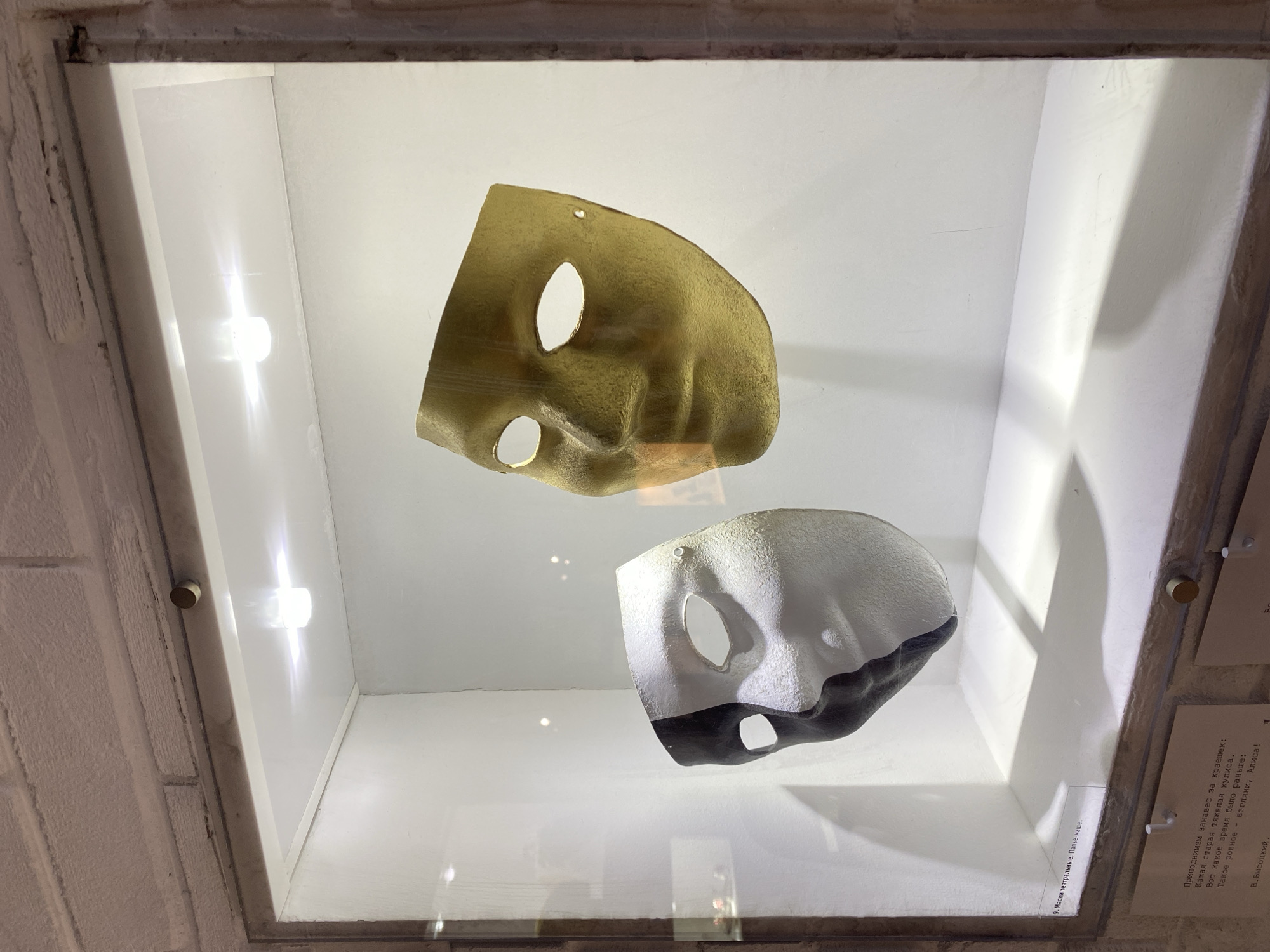 The image shows two theatrical masks displayed in a glass-fronted exhibit case. The mask on the left is split down the middle, with one half painted black and the other half white. It appears to be made of a textured material. The mask on the right is a single, solid color, appearing to be gold or brass. It has a smooth, polished finish. Both masks have openings for eyes and a nose. The exhibit case is lit from above by two bright lights, casting shadows on the white interior walls and the shelf below. A brick wall is visible above the exhibit case. Below the exhibit, there are several pieces of paper with text, suggesting this is part of a museum or gallery display. One of the papers has a label that reads "9. Маски театральные. Папье-маше." which translates to "9. Theatrical masks. Papier-mâché." Based on contextual information, this exhibit is located at the Vysotsky Business Center in Yekaterinburg, Russia, specifically in the museum of Vysotsky.