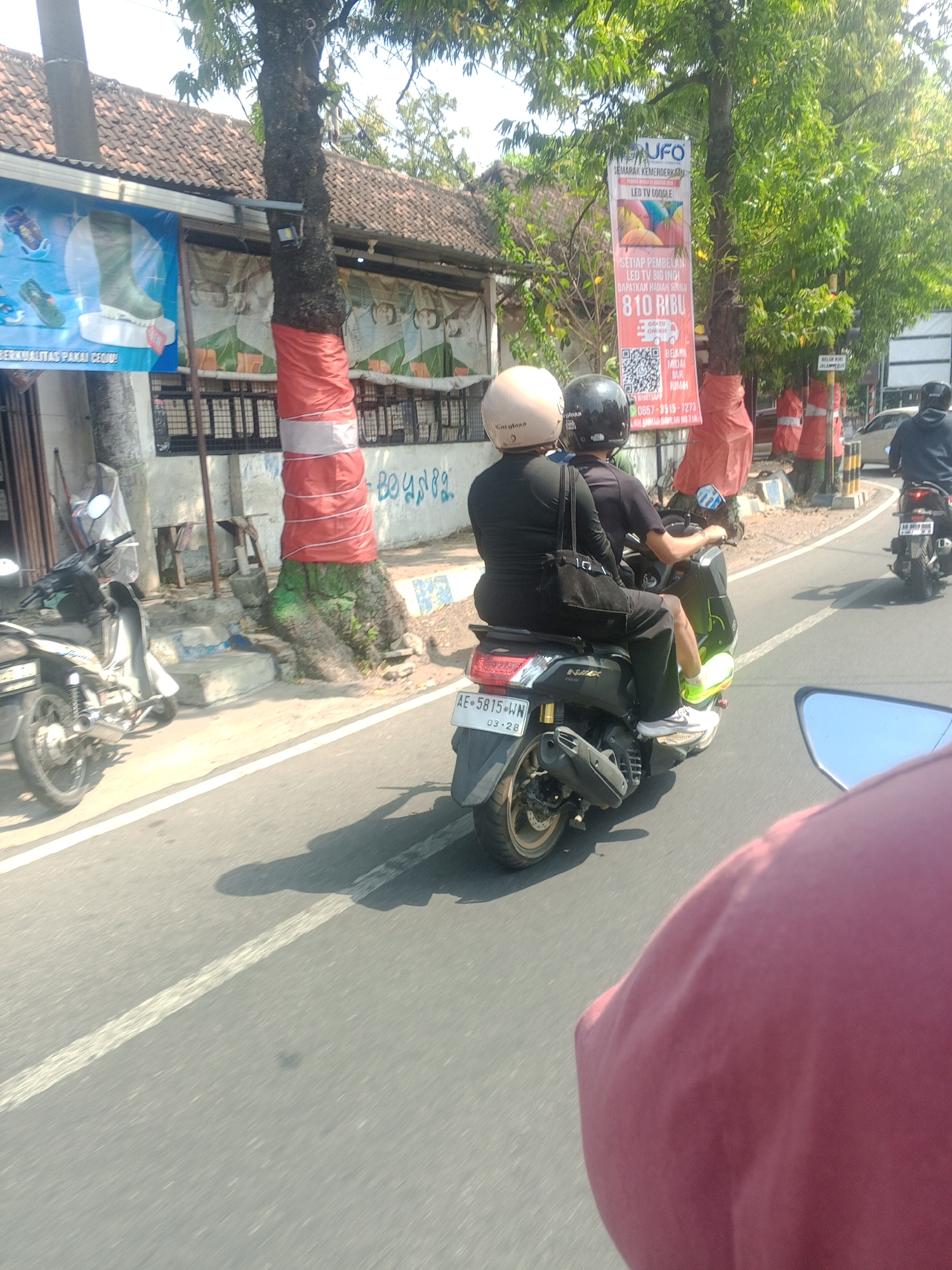 On a sunny day in Nologaten, Indonesia, two individuals are riding a scooter down a paved road. The driver, wearing a black shirt and pants, sports bright yellow sneakers. The passenger, also in black, has a light-colored helmet. A black bag is slung over the passenger's shoulder. The scooter has the license plate AE 5815 WN. In the background, a building with a tiled roof is visible, adorned with red and white banners. One banner displays colorful balloons and text in Indonesian, advertising an LED TV. Trees line the street, one of which is wrapped in red and white fabric. Other scooters and a car are also present on the road. The perspective suggests the photo was taken from another moving vehicle.