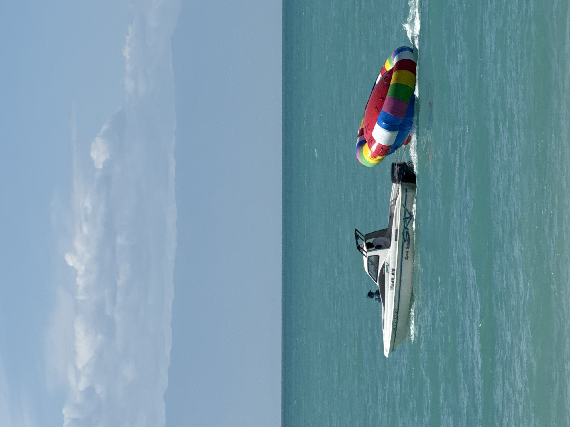 In Zharbulak, Kazakhstan, on a bright day with a clear blue sky and scattered white clouds, a white speedboat with a black canopy is moving across the turquoise waters of a large body of water. Two individuals are visible inside the boat; one is in the driver's seat, and another is seated behind them. The speedboat is towing a large, inflatable towable tube, which is brightly colored with red, yellow, blue, green, and white stripes. The towable tube is buoyant and is creating white wakes in the water as it is pulled by the boat. The background shows a distant, hazy mountain range under the sky, suggesting a vast, open landscape. The scene depicts recreational activity on the water.
