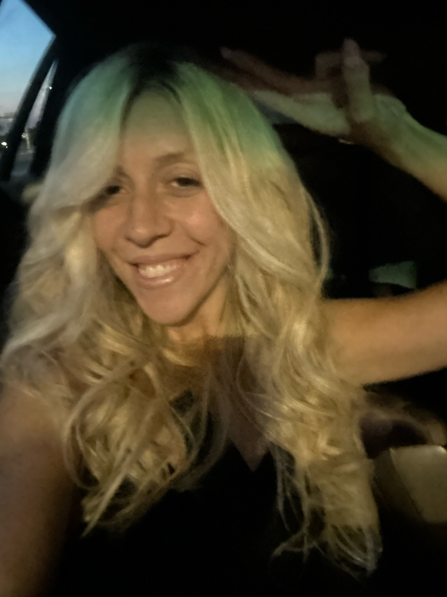 A close-up selfie shows a smiling woman with blonde hair in a car. The lighting suggests it's dusk or nighttime, with a greenish hue cast on her hair. She is wearing a black top. Her right hand is raised, with fingers extended upwards, touching or adjusting her hair. The background is dark, with a faint view of the sky and lights through the car window on the left. There is no identifiable information that can confirm the location as Sofia, Bulgaria.