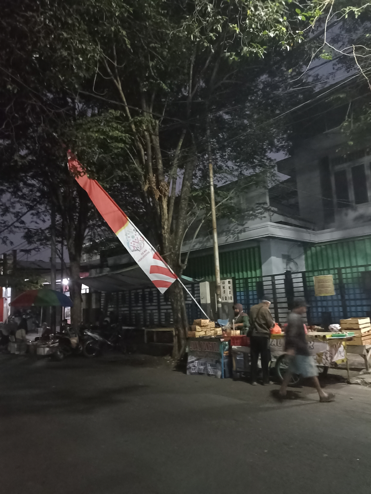A street scene at dusk or night in Ditotrunan, Indonesia, features a red and white Indonesian flag diagonally draped from a tree branch. In the background, a building with a light-colored facade and green slatted doors is visible. In front of the building, several street vendors are set up. One stall has a colorful umbrella, and several people are gathered around. Other vendors display goods on tables and carts, with stacks of what appear to be food items. A few motorcycles are parked nearby, and a person in a grey top and light-colored shorts is walking across the foreground. The street itself is paved and appears empty in the immediate foreground. The lighting suggests artificial streetlights or lights from the vendors' stalls illuminating the scene.