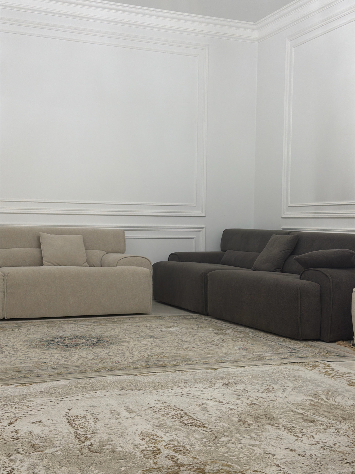 The image depicts an interior scene, likely a living room, featuring two modern sofas and a rug. The room has white walls with decorative molding along the top and framed panels on the walls. A light beige sofa is positioned on the left side of the frame, with a single square cushion on it. To the right, a darker charcoal gray sofa is visible, with multiple cushions. A patterned rug with intricate designs in muted tones of beige, cream, and blue covers the floor in front of the sofas. The lighting appears to be soft and diffused, creating a calm and sophisticated atmosphere. There are no people or active events captured in the image. The setting suggests a residential or a high-end furniture showroom space in Pobedinskoye, Russia.
