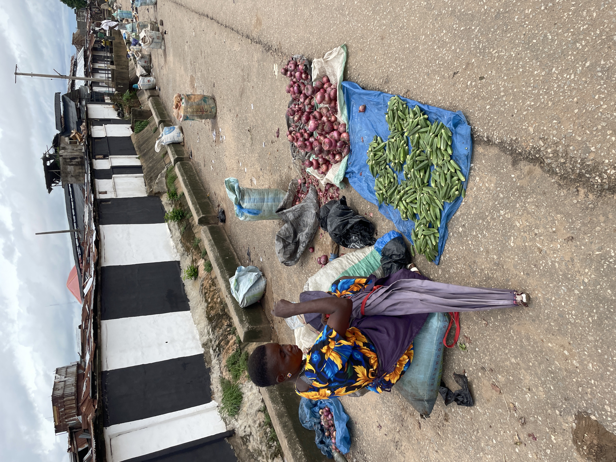 A woman sits on a large sack on the side of a road in an urban environment, surrounded by produce for sale. She is wearing a brightly colored floral top and a purple apron. To her right, there are large piles of red onions and okra spread out on blue tarps and sacks. Behind her, various sacks and bags, some appearing to contain produce or goods, are scattered on the pavement. In the background, there is a row of buildings with a distinctive black and white striped facade, and further down the road, more buildings and some people are visible. The sky is overcast, and the overall scene suggests a local market or roadside vendor setting.