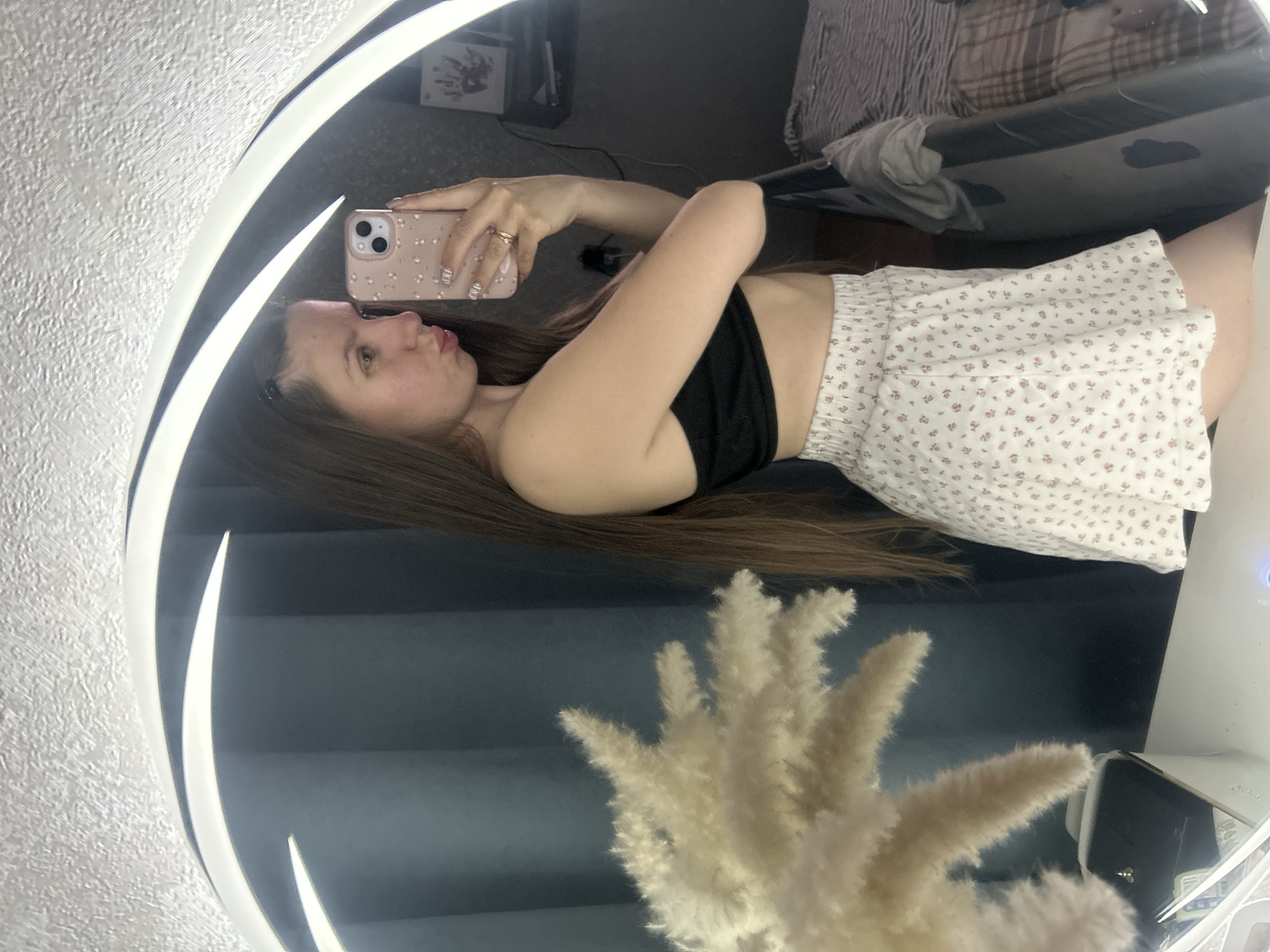 A young woman is taking a selfie in a mirror. She is wearing a black crop top and a white floral skirt. The background includes a bed, blinds, and a decorative plant. There is no discernible information to confirm the location is Klin, Russia.