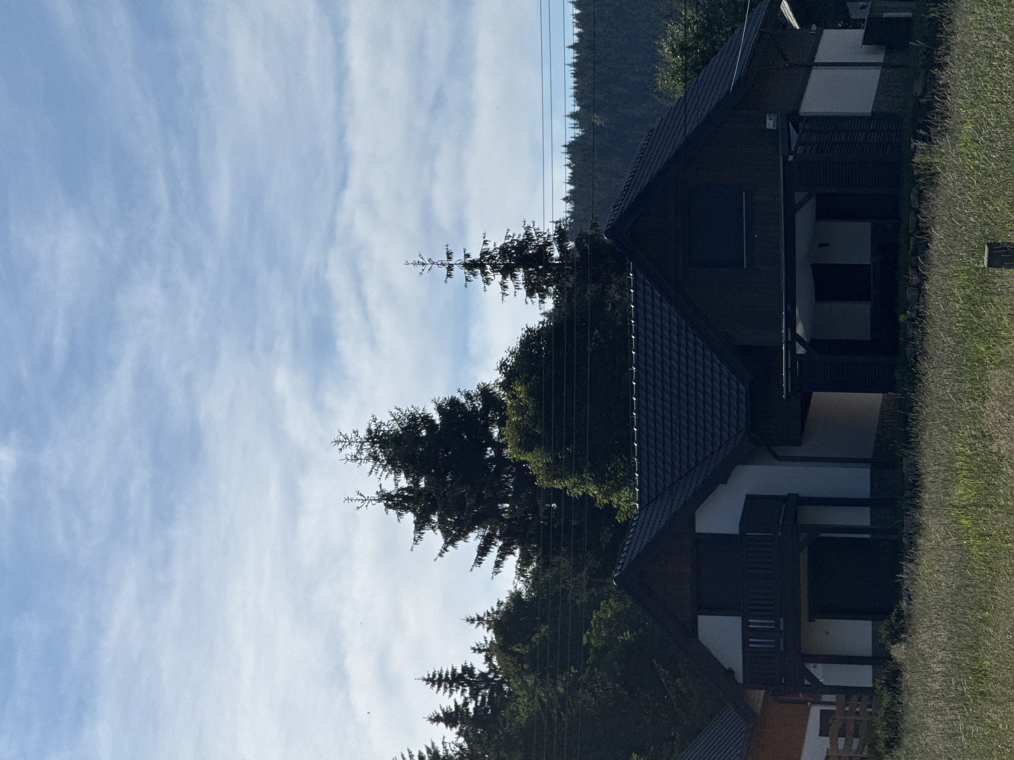 A clear, bright, daytime, outdoor, eye-level shot shows a suburban area. In the foreground, tall green grass grows. In the middle ground, a dark-roofed, multi-story house with white and dark wood siding dominates the scene. The house has a balcony with a wooden railing and an attached covered carport. Behind the house, tall evergreen trees form a dense forest. The sky above is blue with scattered white clouds. Overhead power lines are visible stretching across the frame. The image appears to be taken in Szklarska Poręba, Poland.