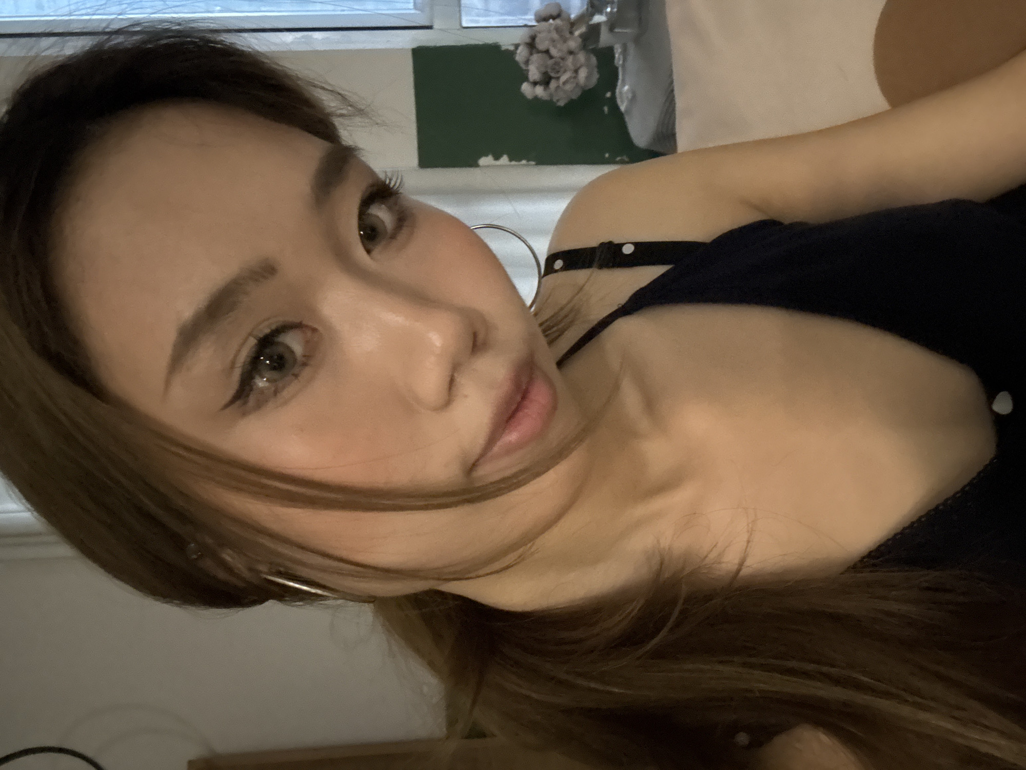 A close-up portrait features a young woman lying on her side, looking directly at the camera, in what appears to be an indoor setting in Liangjiang, China. Her head is tilted at an angle, with her long, dark brown hair cascading across her shoulder and arm. She has clear skin, and her light brown or hazel eyes, enhanced with eyeliner and prominent eyelashes, are the focal point. She wears subtle blush and her lips are slightly parted.
She is dressed in a dark-colored top, possibly a camisole or dress, with a shoulder strap adorned with small white polka dots. A large silver hoop earring is visible on her left ear. In the background, a white vase holds a bunch of light-colored, dried flowers. Below the flowers, a dark green or teal rectangular shape suggests wall decor or a painted accent. A window or diffused light source in the upper background provides soft illumination. The overall mood is calm, with the woman's direct gaze creating an engaging portrait. There is no visible text in the image.