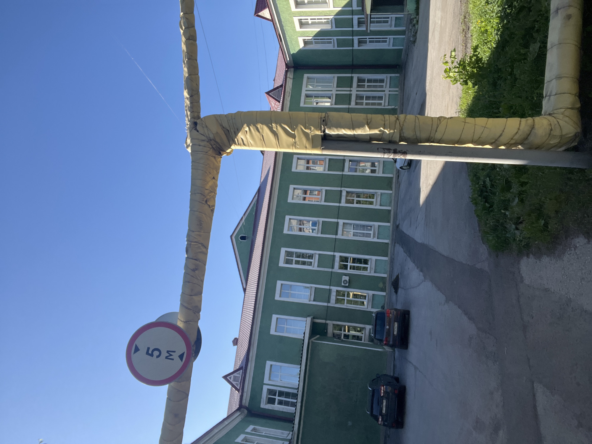 In the city of Kiselyovsk, Russia, a clear, sunny day presents a view of a large, green, two-story building with a red roof. The building features numerous white-framed windows. In the foreground, a large, yellow-insulated pipe runs horizontally across the frame, supported by a vertical pipe. A traffic sign indicating a height restriction of 5 meters is attached to the horizontal pipe. Two dark-colored cars are parked on a paved area in front of the building. The environment is an outdoor urban setting with a bright blue sky overhead.